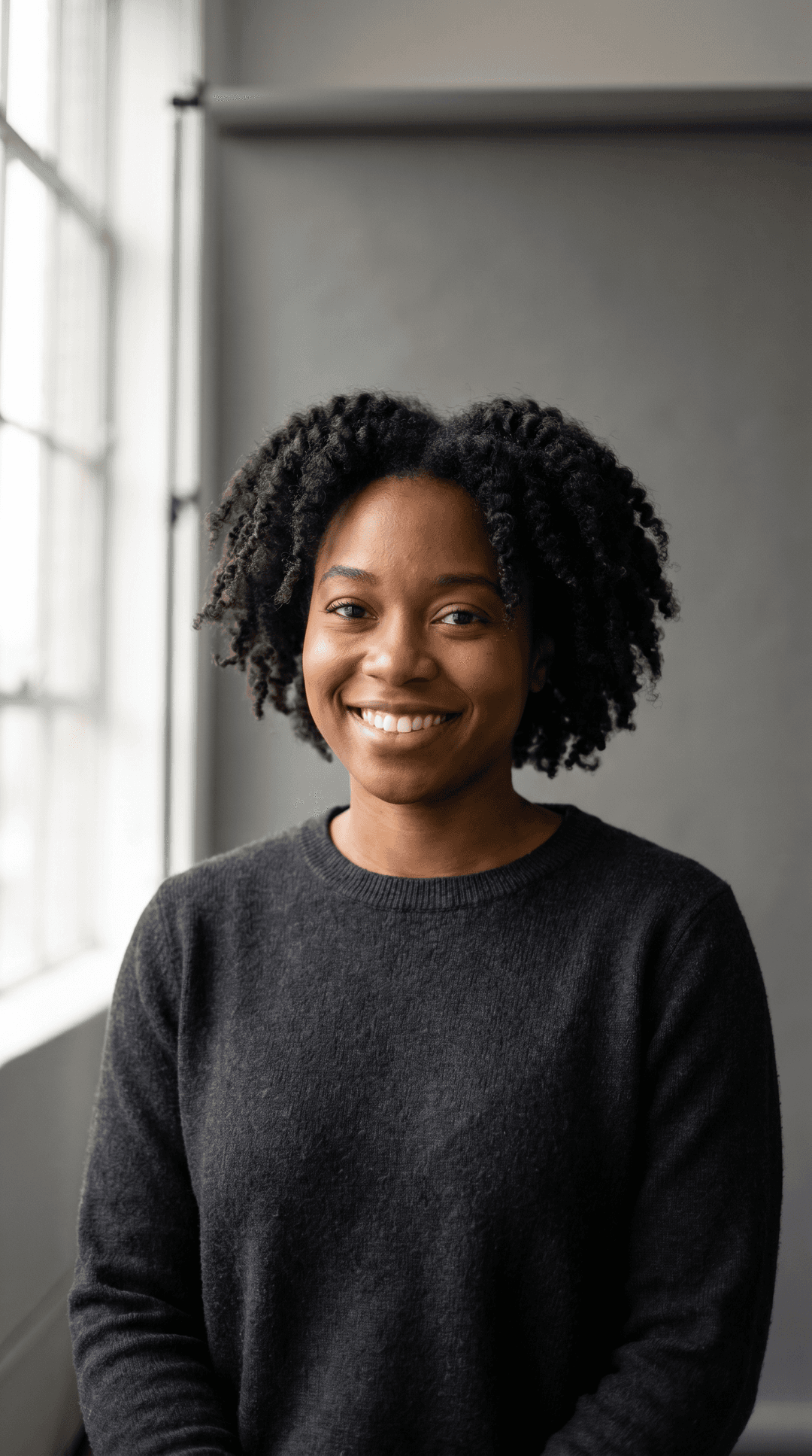 Studio headshot of a friendly Black woman in her late 20s, natural hair, wearing a plain charcoal crewneck sweater. Soft window light from camera-left, neutral grey backdrop, eyes to camera, gentle smile, sharp focus, photoreal, 50mm lens depth of field.