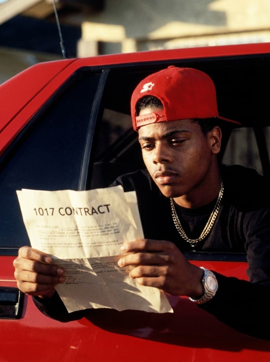 Use the provided image as the base reference. Perform a high-detail character transformation while preserving the original composition.  STRICT RULES: Keep the same camera angle, framing, lighting, and cinematic tone Maintain the same pose and position inside the car window Keep the red car, background, and environment identical Preserve clothing structure (black shirt, red cap) unless naturally adjusted for realism Maintain gritty, street-style cinematic realism  Character Change: Replace the subject with RAPPER Pooh Shiesty  Add subtle jewelry (chain) for realism  Action Change: REMOVE the weapon completely Replace it with the character holding a document/paper in both hands  Document Details: The paper should clearly read: “1017 CONTRACT” Make it look realistic (slightly wrinkled paper, natural grip, proper lighting/shadows) Position it where the weapon originally was for natural composition  Style & Quality: Ultra-realistic, cinematic film still 35mm film look, warm tones, high contrast Natural shadows and highlights Slight film grain for authenticity No cartoon or AI distortion