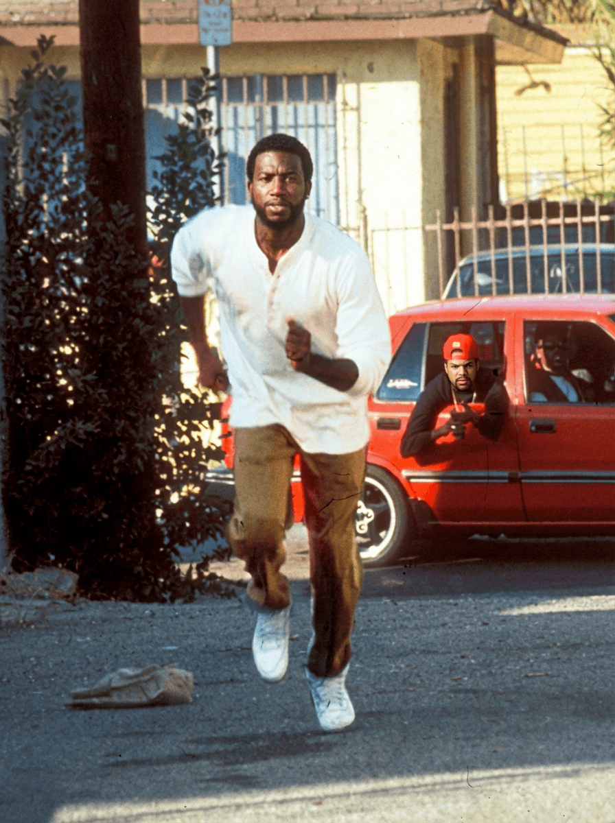 Use Image 1 as the base scene and composition.

🎯 CORE INSTRUCTIONS:

Perform a high-precision dual character replacement while preserving the original cinematic scene.

🏃‍♂️ SUBJECT 1 (RUNNING MAN):
Replace the running man with a realistic version of Gucci Mane (rapper)
Maintain:
Same running pose, stride, and motion blur
Same outfit (white shirt, pants, shoes)
Same body positioning and perspective
Apply:
Gucci Mane facial features, skin tone, beard, and build
Natural expression (focused, mid-run intensity)

⚠️ IMPORTANT RULES:
DO NOT change environment, lighting, or background
DO NOT change the red car or street setting
Maintain original 90s cinematic film aesthetic
Keep natural shadows, highlights, and color grading consistent
Ensure both characters blend seamlessly with correct depth and perspective
🎬 STYLE:
Gritty 90s film still
Slight film grain
Warm tones, high contrast
Realistic motion blur on running subject
No AI distortion or cartoon effects