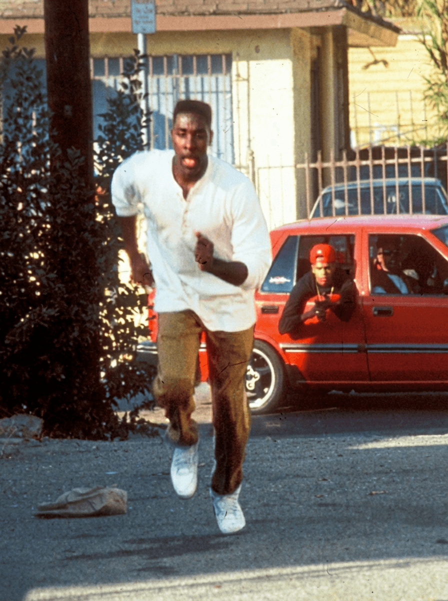 Use the provided image as the base reference.

🎯 CORE INSTRUCTIONS:

Perform a high-precision character transformation while preserving the full cinematic scene.

🏃‍♂️ SUBJECT 1 (RUNNING MAN):
Replace the running man with a realistic version of Gucci Mane (rapper)
Maintain:
Same running pose, stride, and motion blur
Same outfit (white shirt, pants, shoes)
Same body proportions and perspective
Apply:
Gucci Mane facial features, beard, skin tone, and build
Focused, intense expression mid-run

Expression: intense, focused
⚠️ STRICT RULES:
DO NOT change:
Environment, street, or background
Red car details
Camera angle, framing, or composition
Preserve:
Original 90s cinematic look
Natural lighting and shadows
Realistic depth and perspective
🎬 STYLE:
Gritty 90s film still
Slight film grain
Warm tones, high contrast
Natural motion blur on running subject
No cartoon or AI distortion