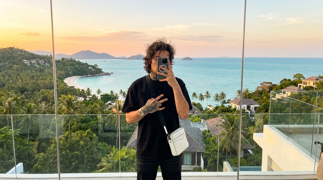 Ultra-realistic, hyper-detailed photograph, captured on an iPhone 16/17 front camera, reflecting in a full-body modern glass mirror. The subject is a young man, centered in the reflection, precisely posed: holding an iPhone obscuring his face, a subtly relaxed posture, one hand forming a delicate gesture near his chest. He wears an oversized black t-shirt, black pants, black shoes, and a white crossbody bag worn diagonally across his torso. His hand and forearm tattoos are exceptionally detailed, sharp, and realistic. His medium-length hair is slightly messy, wavy, and boasts a natural texture.

The setting is a high-end luxury villa balcony on a Koh Samui hilltop. The reflection in the mirror is perfectly proportioned, devoid of distortion.

The background is a breathtaking panoramic vista of Koh Samui: lush tropical jungle, dense palm trees, and steep hills descending into a vibrant turquoise ocean. Multiple small, distant islands dot the horizon. A curved white-sand beach with crystal-clear water stretches along the coastline. Luxury villas are scattered across the hills below, adding to the opulent scene.

The scene is bathed in golden hour lighting, with soft sunset hues – subtle orange and pink tones blending seamlessly into a deep blue sky. Natural light softly illuminates the subject from the front, casting delicate shadows and rendering realistic skin tones with micro skin texture, including visible pores and natural imperfections. Slight warm environmental tones are reflected throughout the image.

The balcony features a modern glass railing with sleek metallic edges, adhering to a minimalistic luxury design. The floor is immaculately tiled. Subtle reflections shimmer on the glass railing, emphasizing the depth perspective of the hill's dramatic drop-off.

The photograph exhibits ultra-sharp focus on the subject, with a subtly softened depth in the background, mimicking natural lens distortion characteristic of a phone camera. High Dynamic Range (HDR) processing ensures extremely realistic lighting and textures.

Enhanced realism is achieved through subtle reflections on the phone and bag materials. A gentle breeze subtly affects the subject's hair. A delicate atmospheric haze hangs over the distant islands. Tiny birds soar or boats glide subtly in the distance, adding a touch of life. Light glints and shimmers on the ocean surface.

The overall mood is one of luxury, calm, and an elevated lifestyle – clean, confident, and effortless. It embodies a perfect fusion of tropical paradise and modern aesthetic.