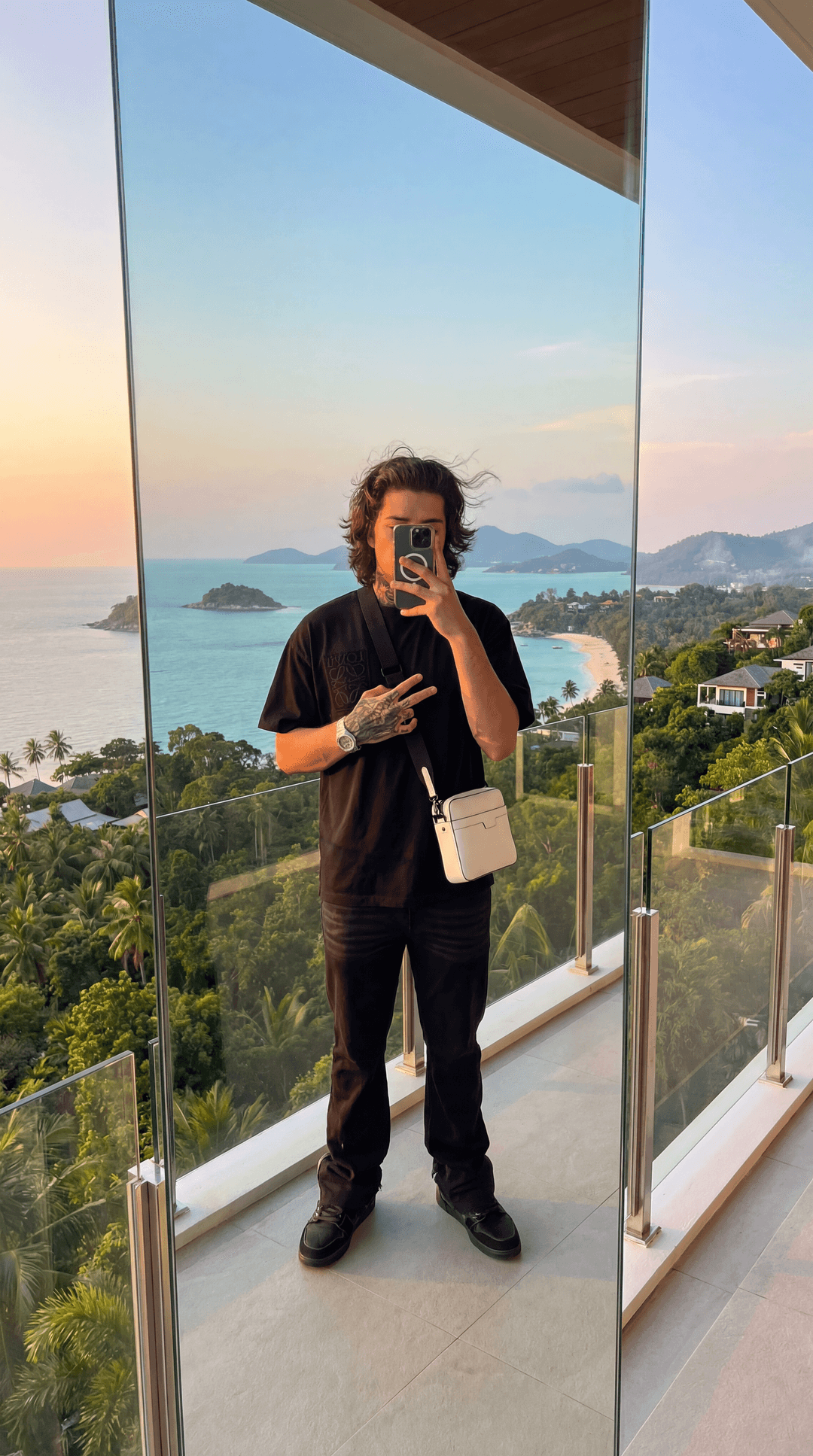 Ultra-realistic photograph of a young man taking a mirror selfie on a luxury balcony located on top of the hills in Koh Samui, Thailand. The subject is standing in the exact same pose: holding an iPhone in front of his face (covering it), slightly relaxed posture, one hand forming a subtle gesture near his chest. His outfit is identical: oversized black t-shirt, black pants, black shoes, and a white crossbody bag worn diagonally across his torso. His tattoos are clearly visible on his hand and forearm, highly detailed and realistic. Hair is medium-length, slightly messy/wavy, natural texture.
The mirror is a full-body modern glass mirror placed on a high-end balcony with a clean luxury villa aesthetic. The reflection shows the subject perfectly centered, with natural proportions and no distortion.
Environment:
The background is a breathtaking panoramic view of Koh Samui from a hilltop villa — lush tropical jungle, palm trees, and steep hills descending into a vibrant turquoise ocean. Multiple small islands visible in the distance. A curved beach coastline with white sand and crystal-clear water. Luxury villas scattered across the hills below.
Lighting:
Golden hour or soft sunset lighting OR optional bright tropical daylight version. Natural light hitting the subject from the front, soft shadows, realistic skin tones. Slight warm tones reflecting from the environment. If sunset: subtle orange/pink tones in the sky blending into blue.
Balcony Details:
Modern glass railing with metallic edges, minimalistic luxury design. Clean tiled floor. Slight reflections on the glass railing. Depth perspective visible with the drop-off from the hill.
Camera Style:
Shot as if taken from an iPhone 16/17 front camera through a mirror. Ultra sharp focus on subject, slightly softened depth in the background. Natural lens distortion consistent with a phone camera. High dynamic range (HDR), extremely realistic lighting and textures.
Extra Realism Enhancers:
Micro skin texture visible (pores, natural imperfections)
Subtle reflections on phone and bag material
Wind slightly affecting hair
Atmospheric haze over distant islands
Birds or boats subtly in the distance for life
Light reflections on the ocean surface
Mood/Vibe:
Luxury, calm, elevated lifestyle. Clean, confident, effortless. Tropical paradise meets modern aesthetic.