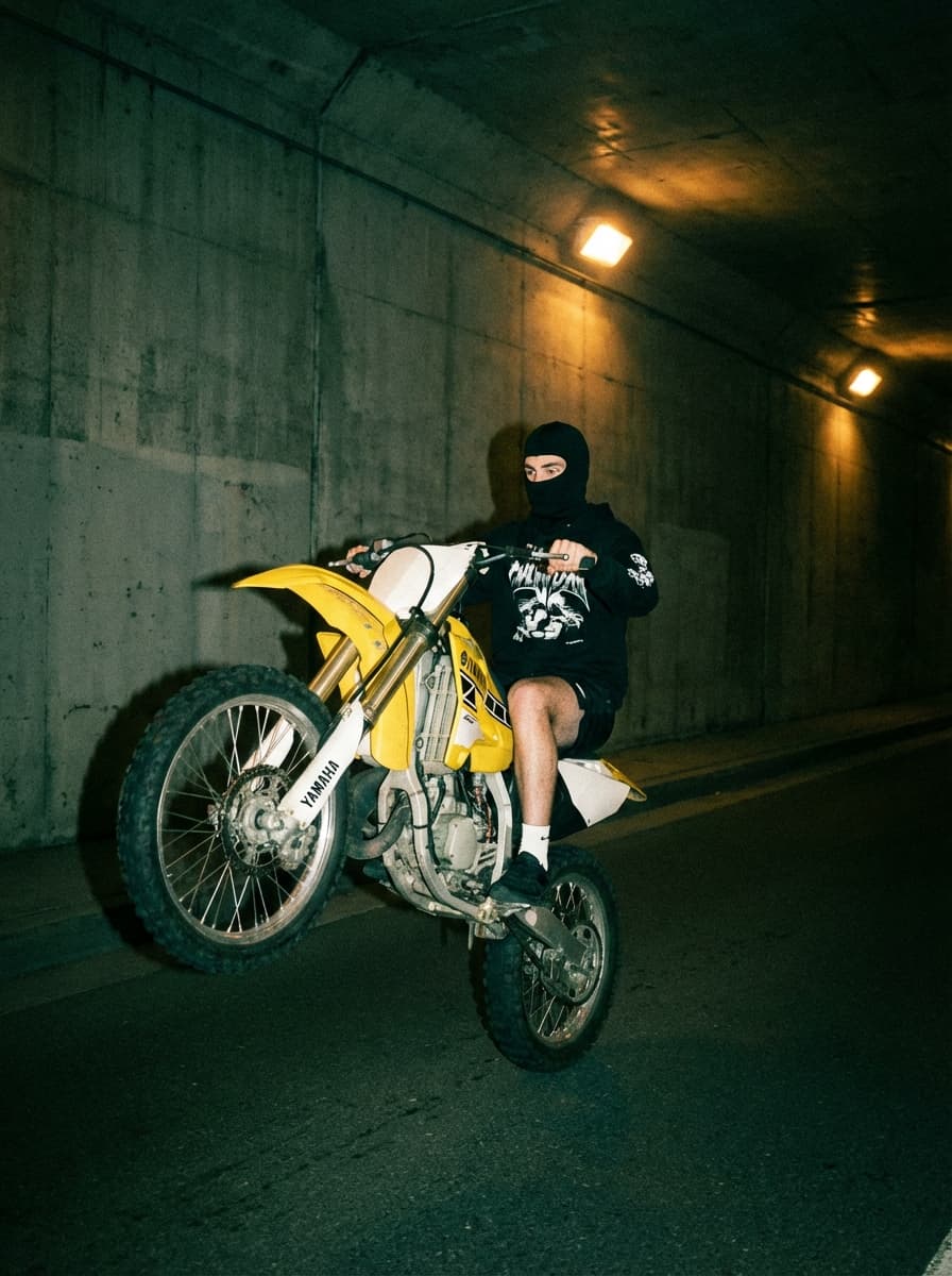 Ultra-realistic nighttime direct flash photography with analog film grain and color-shifted indoor rendering.

Shot on a 35mm film camera (Contax T2 / point-and-shoot film equivalent), 35–38mm lens, aperture f/2.8–f/4, shutter speed 1/60–1/125, ISO 800–1600 film.

Direct on-camera flash with no diffusion. Flash is strong, frontal, and slightly harsh, fully illuminating the foreground while ambient light remains very low.

Lighting behavior:
- Hard shadows with slight softness from film response
- Minimal light falloff beyond flash range
- Background fades into darkness with faint ambient visibility

Highlights are bright but softer than digital — slight bloom and gentle roll-off typical of film. No harsh digital clipping, but still punchy.

Color grading features a green/teal cast in shadows with warm highlights, creating a mixed-color, slightly contaminated film look. Whites are not pure — slightly tinted and aged.

Contrast curve:
- Blacks moderately crushed but still retain some texture
- Midtones slightly compressed
- Highlights softly rolled off with mild glow

Saturation is muted:
- Colors slightly desaturated and shifted
- Overall palette feels subdued and imperfect

Color profile resembles pushed 35mm film (Fuji Superia / Portra 800 pushed) with color imbalance and analog inconsistency.

Grain is prominent and organic:
- Visible 35mm film grain across entire image
- Soft, clumpy grain structure (not digital noise)

Sharpness is moderate:
- Slight softness from lens + film
- Reduced edge sharpness compared to digital

Dynamic range is moderate with soft highlight handling and slightly lifted shadows compared to digital flash.

Framing is medium-wide with natural eye-level perspective (35–38mm). Slight handheld looseness with documentary feel.

Depth of field is shallow to moderate — subject sharp, background slightly soft.

No motion blur, but overall image retains a softer analog feel.

Overall aesthetic: vintage film flash / late-night documentary / analog snapshot realism.

Maintain film grain, green/teal shadow cast, soft highlight bloom, and imperfect color balance. Do not clean or modernize the image.

SUBJECT + ENVIRONMENT:  Ultra-realistic photograph.

Young adult male with light-medium skin tone, athletic build. Face mostly covered by a black balaclava exposing only eyes and upper nose. Eyes focused forward with alert intensity. Wearing a black long-sleeve hoodie with bold white abstract graphic patterns across sleeves and chest, black shorts, black gloves, white crew socks, and white low-top sneakers. Legs exposed from mid-thigh down showing natural skin texture.

Riding a yellow Yamaha dirt bike in a controlled wheelie position. Front wheel lifted high off the ground at a steep upward angle, rear wheel maintaining contact with the asphalt. Rider leaning slightly backward with hips seated on the rear portion of the bike, arms extended forward gripping handlebars, left foot planted on foot peg, right foot positioned near rear brake. Body balanced precisely to maintain wheelie.

Urban tunnel or underpass environment at night. Background consists of large concrete wall panels with evenly spaced circular indentations and visible seams. Upper portion of frame shows warm overhead industrial lighting casting orange glow onto concrete. Ground is dark asphalt road with subtle texture and faint reflective sheen.

Shot on Canon EOS 5D Mark IV, 28mm lens, aperture f/4, shutter 1/200, ISO 800.

Lighting: direct on-camera flash as primary light source combined with ambient tunnel lighting. Flash produces harsh frontal illumination with strong specular highlights on bike surfaces and clothing. Hard-edged shadows cast sharply onto the concrete wall behind subject. Ambient sodium-vapor style lighting adds warm orange cast in upper background, mixing with neutral flash foreground.

Color science: mixed white balance with cool-neutral flash foreground and warm orange ambient highlights in background. High contrast with slightly crushed blacks. Highlights bright with slight clipping on reflective metal parts of bike. Yellow bike remains vivid and saturated. Skin tones neutral but slightly flattened by flash. Fine digital grain present consistent with mid ISO.

Composition: medium-wide shot with slight wide-angle perspective. Camera positioned at low waist height angled slightly upward toward subject. Subject centered slightly right, bike diagonally oriented upward creating dynamic vertical motion. Tight framing with minimal negative space above wheel. Perspective exaggerates front wheel height and depth.

Depth: moderate depth of field with rider and bike in sharp focus. Background wall slightly softened but still clearly defined. Separation achieved through flash contrast and subject proximity rather than heavy blur.