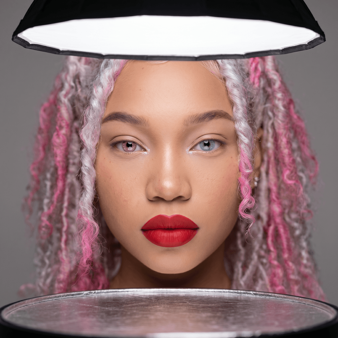 A close-up, high-fashion, studio portrait of a woman with striking, symmetrical features, shot with a Canon EOS R5 and a RF 85mm f/1.2L USM lens. The lighting is a classic clamshell setup with a large parabolic softbox above and a silver reflector below, creating soft, even illumination and catchlights that emphasize her eyes. Her skin is flawlessly smooth, with subtle natural texture, and her makeup is minimalist but impactful – a bold, matte red lip and perfectly shaped brows, with a hint of shimmer on the inner corners of her eyes. The background is a seamless, neutral grey, providing a clean and uncluttered canvas for her face. The focus is razor-sharp on her eyes, with a shallow depth of field that gently blurs her hair and ears. The overall aesthetic is one of elegant sophistication and timeless beauty, with a hyper-realistic quality that captures every minute detail.