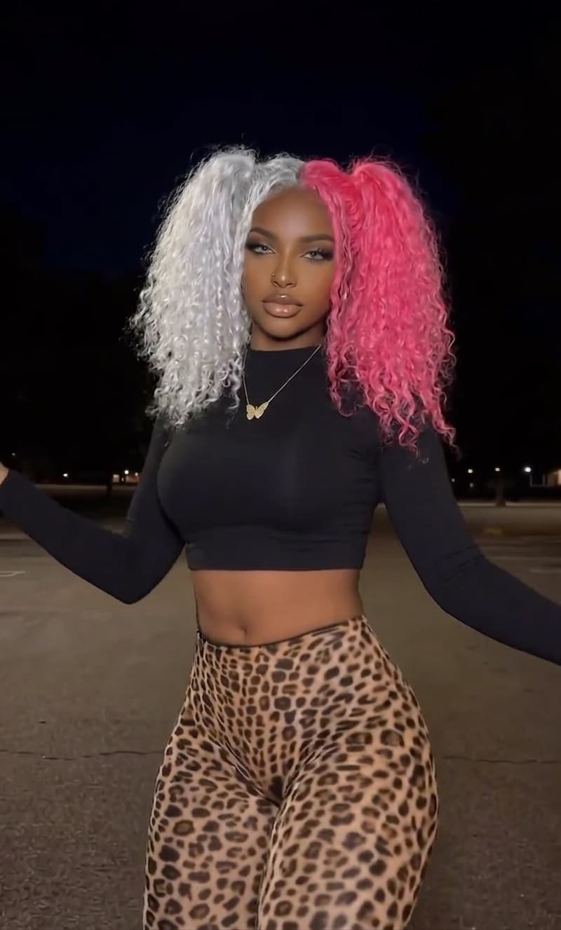 Replace the woman with black curly hair with the same woman who has voluminous curly hair styled in high pigtails — half white/silver, half bright pink. She is wearing the same black long-sleeve crop top and high-waisted, form-fitting leopard-print leggings. She maintains the same confident pose standing with arms slightly outstretched, exuding confidence and poise outdoors at night. The background remains the same dark, empty parking lot or open paved area at night with minimal distractions. The dim ambient lighting creates a dramatic, moody nighttime atmosphere. She has the same polished makeup with defined brows, smoky eyes, glossy lips, nose ring, and delicate gold necklace with pendant. Focus entirely on her striking appearance against the dark background, exactly as the original pose but with the pink and white hair instead of black hair.