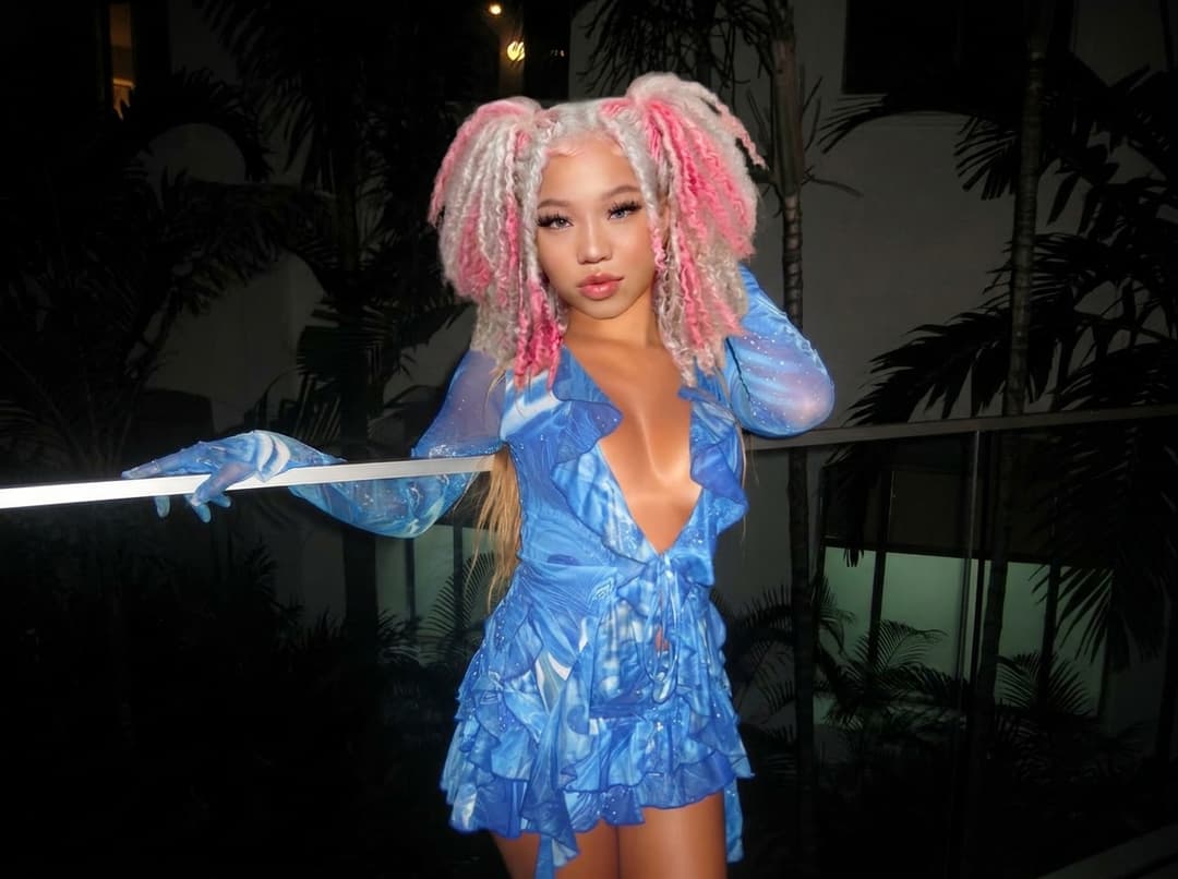 Replace the face, skin tone, and hairstyle of the blonde hair woman with the pink and white hair woman reference person, while keeping the original pose, body, outfit, lighting, and background unchanged. Ensure the result looks natural, realistic, and seamlessly blended