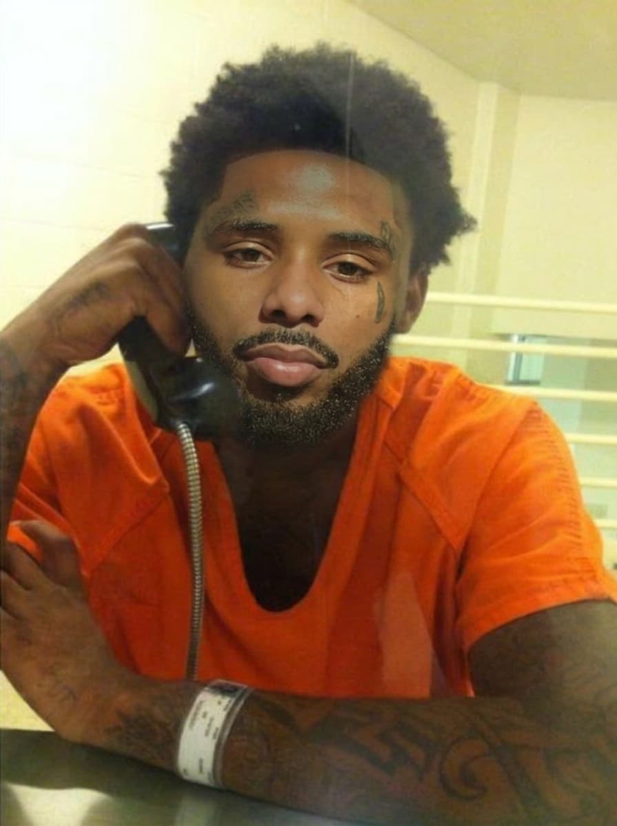 replace the man on phone in jail face with pooh shiesty (refence man) keeping lighting changing nothing else keep reference man facial hair in swap keep lighting and bad quality of jail photo and keep face proportionate