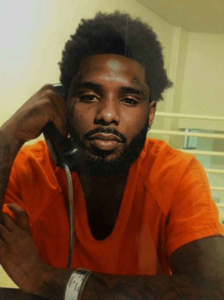 replace the man on phone in jail face with pooh shiesty (refence man) keeping lighting changing nothing else keep reference man facial hair in swap keep lighting and bad quality of jail photo and keep face proportionate