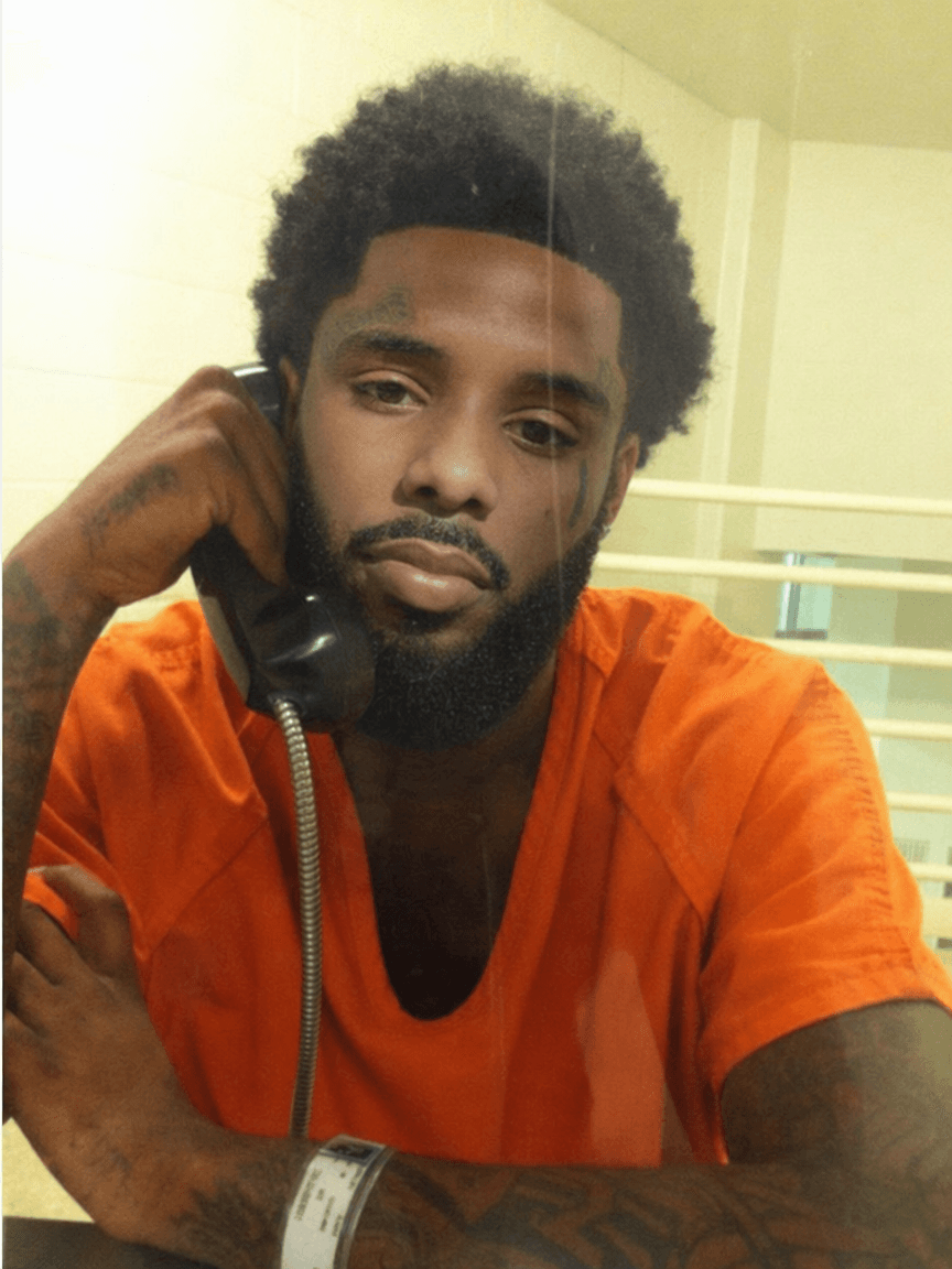 replace the man on phone in jail face with pooh shiesty (refence man) keeping lighting changing nothing else keep reference man facial hair in swap keep lighting and bad quality of jail photo and keep face proportionate