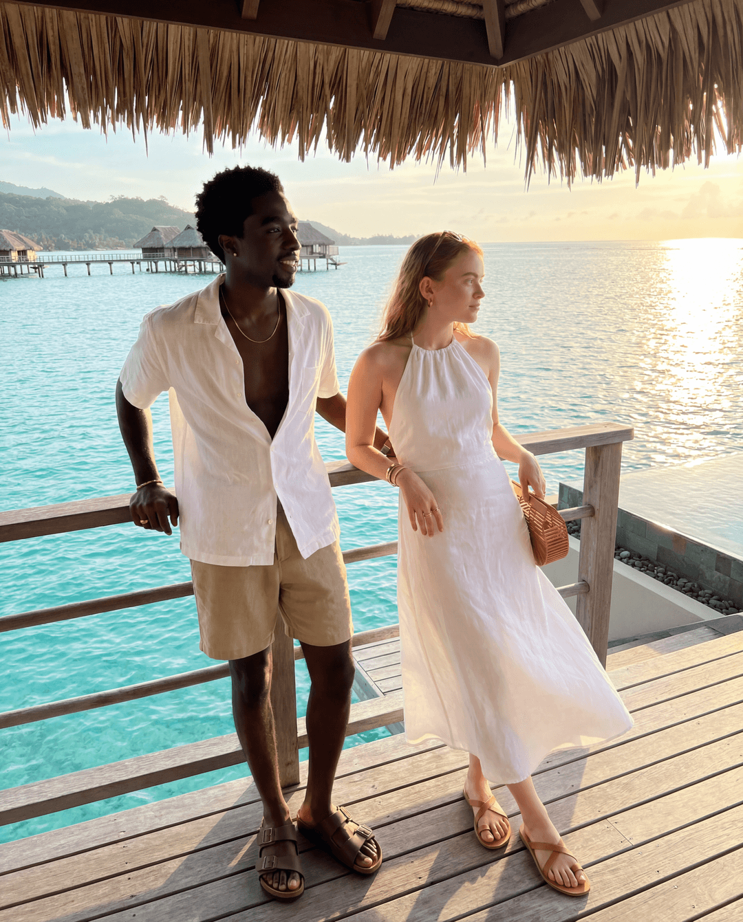 Camera settings: iPhone 17 Pro Max, 48MP sensor, natural bright Bali tropical golden hour sunlight warm and glowing, zero filters, real skin texture pores visible, wide shot full body both standing together on over water bungalow deck, true to life colors, handheld candid friend energy, real tropical water and resort environment visible. Caleb McLaughlin and Sadie Sink standing together on the private deck of a luxury over water bungalow at a Five Star Bali resort, turquoise Indian Ocean water stretching endlessly in every direction below and behind them, natural candid relaxed vacation friend energy not posed. Caleb McLaughlin standing left, one hand resting on wooden deck railing casual, other hand in pocket, looking out over the turquoise ocean natural unbothered vacation energy slight smile, wearing a white linen short sleeve button up shirt open at collar real linen texture slightly wrinkled from Bali humidity, khaki linen shorts real heavy linen texture, brown leather Birkenstock Arizona sandals real worn leather footbed, thin gold chain at neck catching warm golden hour Bali light, no watch just relaxed. Sadie Sink standing right beside him, both arms resting on wooden deck railing leaning forward slightly looking out over ocean, genuine peaceful expression taking in the view, wearing a white Reformation linen halter sundress thin straps real linen texture slightly billowing in warm ocean breeze, tan leather Ancient Greek Sandals flat strappy sandals real leather strap detail, small woven rattan Cult Gaia bag on deck railing beside her, small gold hoop earrings catching tropical golden hour light, Cartier Love bracelet yellow gold stack catching warm light. Luxury over water bungalow deck real teak wood planks underfoot real wood grain texture, wooden railing in front of both real weathered teak detail, turquoise crystal clear Indian Ocean water directly below deck visible through gaps in deck boards real water color depth, other over water bungalows visible in distance on both sides connected by wooden walkways stretching over water, lush Bali jungle covered hills visible on horizon in far distance, golden hour sun low casting everything warm amber and gold, sun path reflecting on ocean surface real golden light shimmer on water, palm trees on resort island real visible left side, traditional Balinese thatched grass roof of bungalow visible above in upper frame, infinity pool edge barely visible at far right of deck, warm golden Bali breeze moving Sadie dress hem and hair naturally, zero AI glow, real teak wood deck texture underfoot, real linen texture both outfits catching warm golden ocean light, sandal leather real, jewelry catching golden Bali sun naturally, skin pores and natural imperfections fully visible both faces in warm tropical golden hour light, ocean water real turquoise color depth below deck, ultra realistic iPhone Bali over water bungalow resort wide vacation candid capture. 
