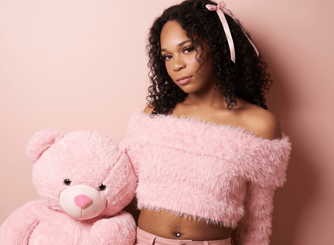 A young woman with long dark hair, wearing a fashionable pink fluffy off-the-shoulder top, posing elegantly in front of a pink wall with a large pink stuffed animal next to her. She has the exact same face as the original mirror selfie girl - same facial features, same expression, same makeup. She has a pink ribbon in her curly dark hair and a belly button piercing visible. High quality fashion photo with professional lighting, Instagram aesthetic. The face must be identical to the original mirror selfie face, just placed on this new fashionable body in a pink setting.