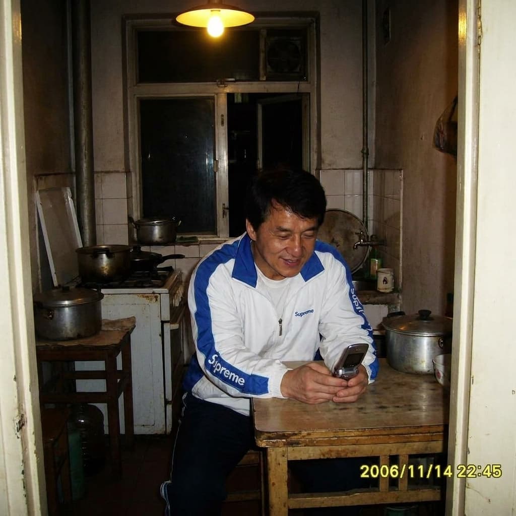 Jackie chan sitting in a small kitchen at night, overhead light on, quiet atmosphere, phone-quality image, almost real wearing a " supreme " white and blue jacket 