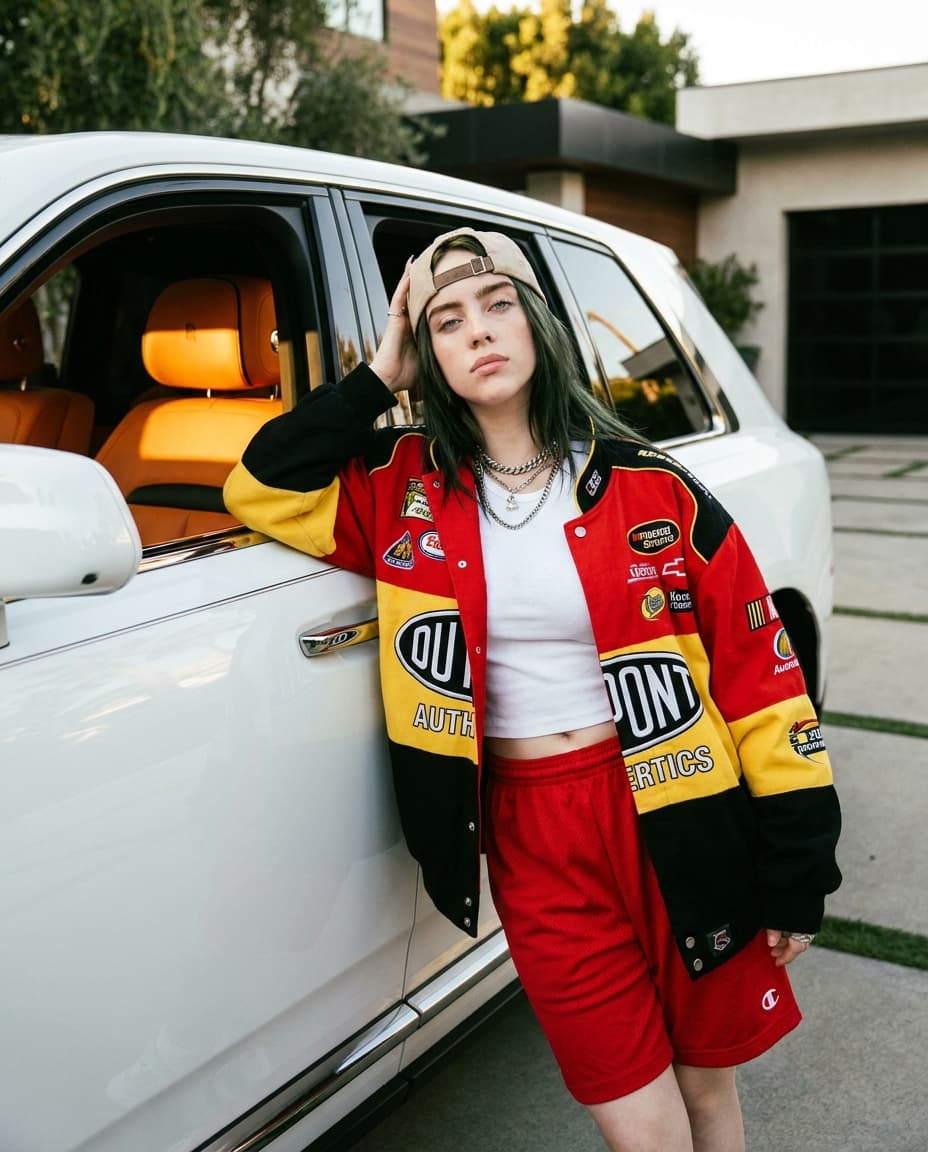 **4K ultra detailed hyper-realistic cinematic celebrity photography of Billie Eilish stepping out of a luxury white Rolls-Royce Cullinan SUV, driver door ajar, during golden hour.**

Billie embodies her signature oversized luxury streetwear aesthetic infused with feminine Y2K glam: an oversized, vibrant red, yellow, and black racing jacket adorned with authentic motorsport patches, a cropped white baby tee, perfectly fitted red athletic shorts, a beige baseball cap worn backward, and multiple layered silver chains. Her long black hair features subtle dark green undertones, framing a face with soft glam makeup. She exudes confident celebrity energy.

**Pose:** Billie leans casually against the open SUV door, one arm gracefully stretched across the door frame, the other resting near her head. One knee is bent slightly forward, conveying relaxed rich-girl body language and an emotionally detached yet stylish expression.

**Details:** The luxurious orange leather interior of the Rolls-Royce glows behind her, casting warm light. Glossy reflections shimmer on the pristine white paintwork of the SUV. Realistic skin textures are meticulously rendered under the natural warm sunlight. The image captures a paparazzi-meets-editorial photography aesthetic, evoking a luxury influencer atmosphere and cinematic realism with a shallow depth of field. Highly detailed fashion styling is evident throughout.

**Background:** An upscale mansion driveway with contemporary architecture and soft, blurred greenery in the distance.

**Style:** Tumblr-core luxury aesthetic, subtle film grain, glossy highlights, realistic shadows, expensive influencer vibe.

**Format:** 4:5 vertical Instagram editorial image.