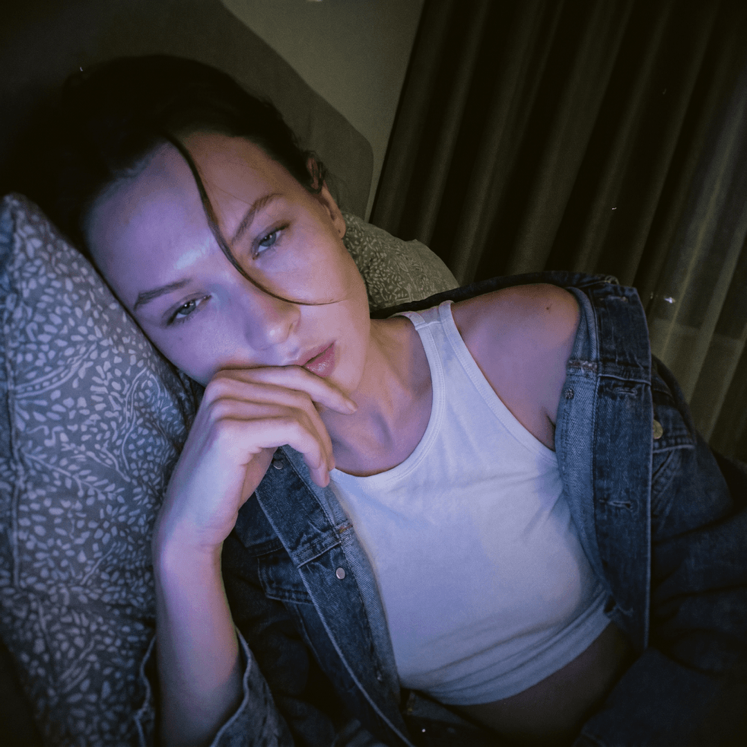 <faceid:3983732:1.0> { "meta": { "aspect_ratio": "9:16", "quality": "ultra_photorealistic", "resolution": "8k", "camera": "iPhone 15 Pro Max front camera", "lens": "24mm wide", "style": "raw iPhone selfie realism, not studio, not professional, visible natural texture" }, "character_lock": { "face_identity": [ "same as reference image", "same facial proportions, same jawline", "NO face change, NO face swap errors" ], "skin": [ "pale porcelain skin tone", "smooth but real texture", "not plastic, not over-smoothed", "soft glow on cheeks" ], "hair": [ " hair", "messy bedroom hair", "side-part with strands falling over one eye", "slightly shiny strands, soft volume" ] }, "scene": { "location": "dim bedroom at night", "environment": [ "dark background", "soft shadows", "beige blackout curtains on left side", "bed vibe, cozy messy room feeling" ], "atmosphere": "late-night sleepy lo-fi, intimate quiet mood" }, "lighting": { "type": "low-light cool glow", "key_light": "cool bluish-purple glow on face", "fill_light": "very soft ambient darkness", "contrast": "high contrast, face lit but background nearly black", ] }, "camera_perspective": { "pov": "arms length selfie", "angle": "slightly low angle close-up selfie", "distance": "very close, intimate framing", "framing": "face + upper chest, cropped tight, arm extended and leaving the frame at the edge", }, "subject": { "gender": "female", "age": "21+ (adult)", "vibe": "effortlessly hot, sleepy, soft but dangerous", "expression": { "eyes": "heavy-lidded dreamy stare", "mouth": "slightly open, relaxed lips", "emotion": "tired + seductive without trying" }, "pose": { "position": "lying on stomach", "support": "propped up on grey pillow with subtle pattern", "hand": "hand near face, index finger touching lower lip" }, "outfit": { "top": { "type": "extremely cropped white tank top / tee", "fit": "skin-tight, stretched thin, riding up high", "details": "thin fabric, realistic tension folds, visible through material", "underwear": "no bra" }, "extra": [ "denim jacket completely slipped off shoulders and down arms, barely hanging on elbows", "black lace thong visible above very low-rise jeans that are unbuttoned and pulled halfway down hips", "jeans showing just the top edge of lace underwear and bare midriff/lower stomach" ] } }, "image_quality": { "focus": "soft focus but face remains clear", "grain": "visible low-light noise", "sharpness": "NOT razor sharp, more lo-fi", "realism": "looks like a real iPhone selfie posted online" } 