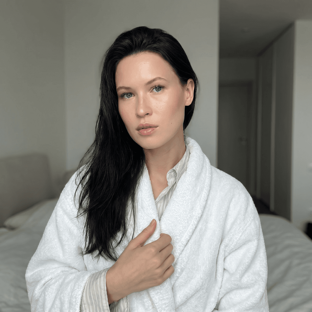 A medium shot captures the person in the uploaded reference image (DO NOT TOUCH FACE) from the chest up, angled slightly to the left. They are wearing a plush, white terrycloth bathrobe, open at the front to reveal the upper chest. Their left arm is bent, with the hand resting near the center of their chest, holding the robe closed. Long, straight, dark hair cascades over their left shoulder and down their back. The background is a softly lit, modern interior, with a blurred view of a bed and a closet door visible on the right. The lighting is soft and diffused, coming from the front and slightly to the left, creating subtle highlights on the skin and fabric. The camera is at eye level, with a shallow depth of field that blurs the background.
