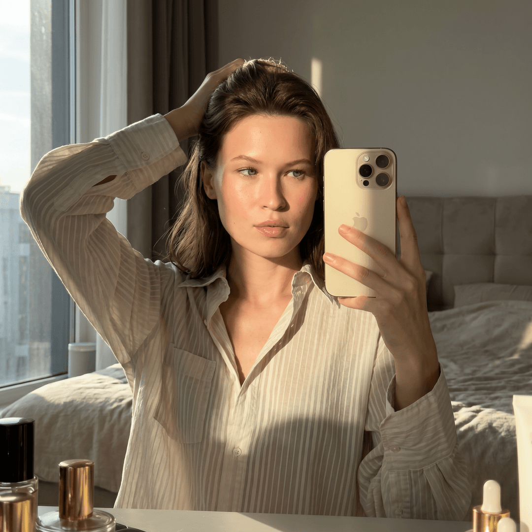 Using the person in the uploaded reference image, recreate this scene: ultra HD photography in the morning. Face matching both 100%. Woman 35s Close-up standing selfie in the mirror, one hand pulling back hair, gold iPhone 17 Pro Max visible in the reflection wearing clothes by reference in second image. Lighting: Golden sunlight through a window, high contrast. Mood: Radiant luxury, sensual realism. Don't replace both of their faces.