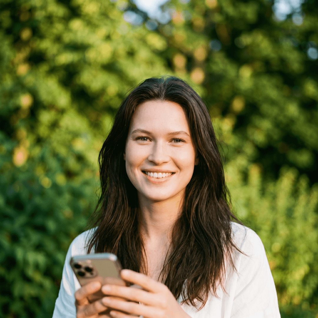 A photorealistic lifestyle portrait of a smiling woman from the uploaded image looking directly at the camera, DO NOT TOUCH FACE. Her head is slightly tilted. She has long dark hair flowing over her shoulders and is wearing a simple white top. Her left hand is gently holding a blurred object, possibly a phone, near her chest. Shot outdoors with soft, natural golden hour light, casting a gentle glow, featuring a shallow depth of field and creamy bokeh in the background with lush green foliage. Eye-level shot, framed from the chest up, close-up. Natural color grading, soft contrast, detailed textures, no over-processing.
