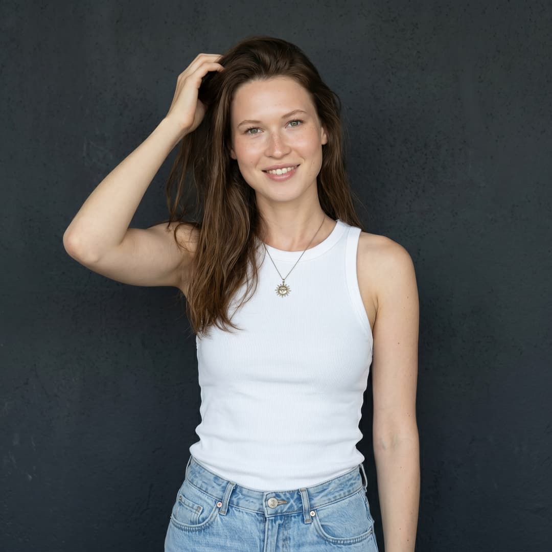 A medium shot captures the person in the uploaded reference image (DO NOT TOUCH FACE) wearing a white ribbed tank top and light wash denim jeans, standing against a dark, textured wall. The person has their hand raised, fingers lightly touching their hair, and is looking off to the side with a slight smile and freckles visible on their face. A delicate sun pendant necklace is worn over the tank top. The lighting is natural and directional, casting soft shadows and highlighting the textures of the clothing and wall.