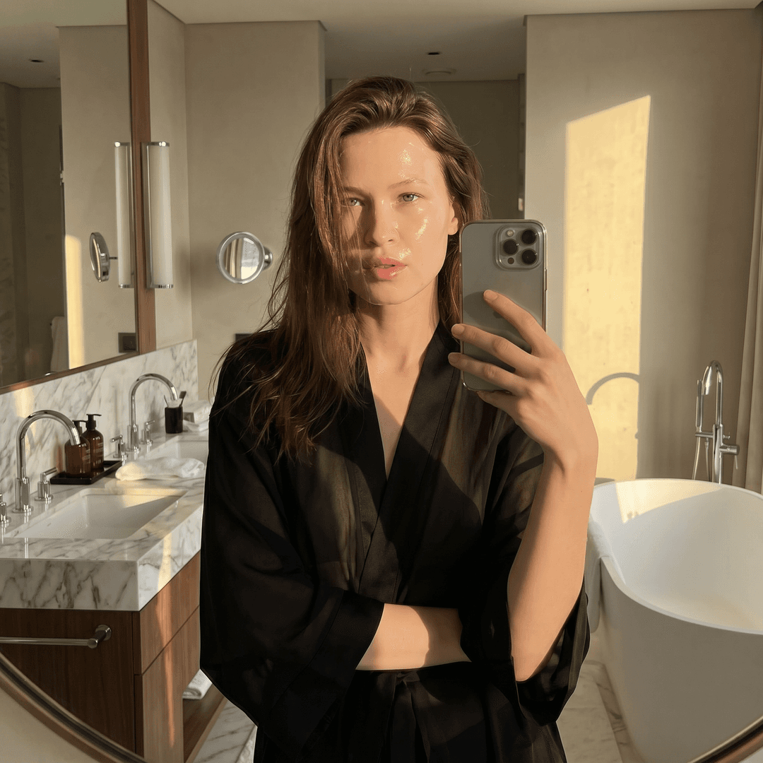 Selfie of the person from the reference image model standing in a bright expensive bathroom. Face with a natural glow. Face half-turned. She is wearing a black просвечивающийся silk robe. On the face is a thin gel care mask. Golden hour lighting, ultra-realistic, aesthetic, high quality. Do not change my real facial features. Shiny hair partially on the face.