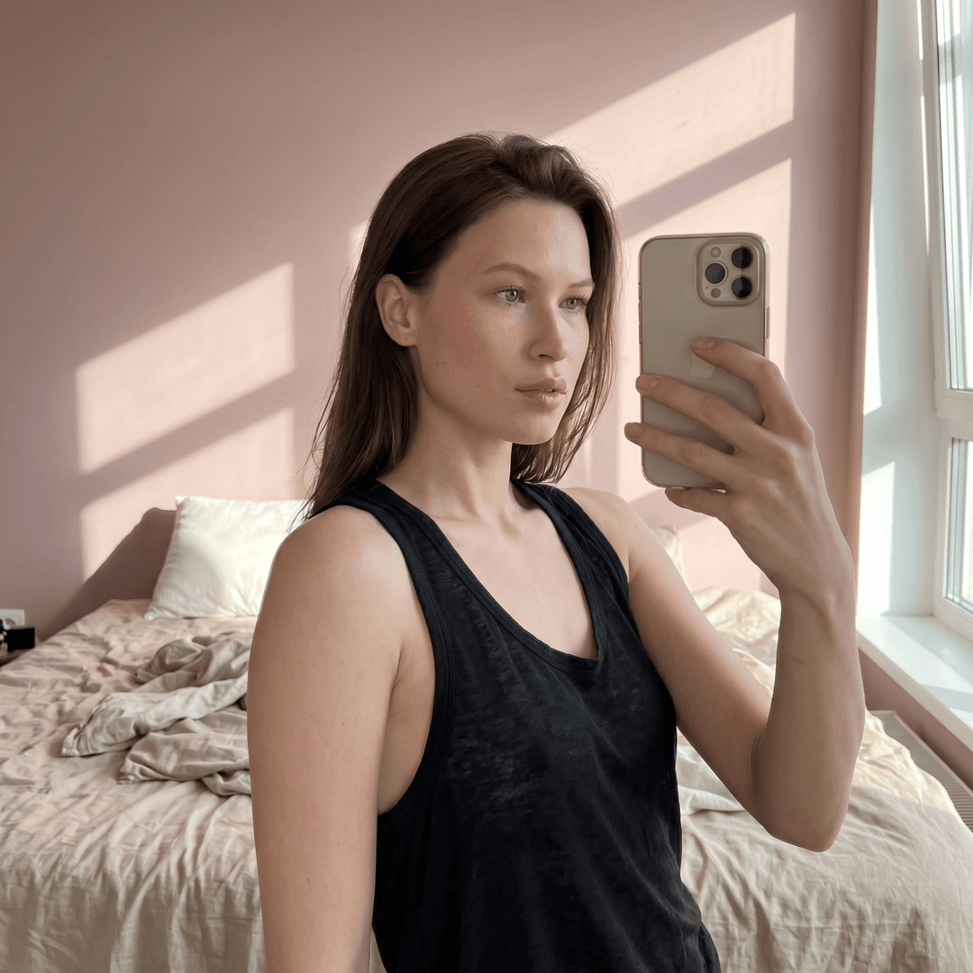 “A scene featuring the woman in the uploaded reference image (DO NOT TOUCH FACE). A medium shot captured by a smartphone held at arm's length, with a shallow depth of field. The subject is positioned slightly off-center to the left, looking towards the right. Their right hand. They wear a black, просвечивающияся, tank top made of a soft cotton blend. майка слегка небрежно растянута. девушка The background is a bedroom with a rumpled bed covered in light beige linens. Sunlight streams in from a window on the right, casting diagonal shadows across the scene. The wall behind is painted a soft pink. The camera is at eye level, with a subtle 24-28mm equivalent lens perspective. The lighting is natural and warm, with highlights on the subject's face and hair. ”


