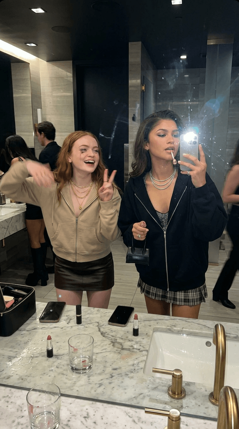 Idea: luxury girls-night-out bathroom photo that accidentally looks like a campaign
Clothing Style: oversized zip hoodies, mini skirts, knee-high boots, layered necklaces, tiny handbags hanging from wrists
Camera Angle: mirror selfie style iPhone 16 Pro flash shot

A hyper-realistic luxury bathroom image featuring the two uploaded subjects taking a chaotic mirror flash photo together inside a fancy nightclub bathroom. One girl fixes her lip gloss while staring at herself instead of the camera. The other aggressively flashes a peace sign while laughing with blurry shoulder movement.

The bathroom should feel expensive but messy:
water droplets on the sink, lipstick marks on cups, chrome reflections, mirror smudges, wet counter reflections.

Flash creates:
hot skin highlights, realistic makeup texture separation, visible pores, authentic skin shine, slight mascara smudging under eyes, natural facial texture. The framing should feel rushed and imperfect like a real girls-night memory.