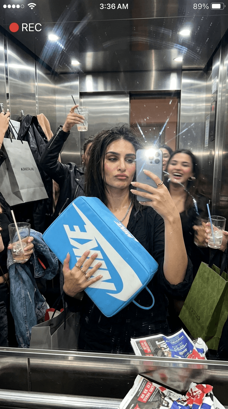 A hyper-realistic, messy glamorous UGC flash photograph, captured as a mirror selfie from an iPhone 16 Pro, inside a cramped, chrome-lined elevator after a night out. The subject, with slightly drunk laughter, sleepy eyes, and a playful pout, holds the PRODUCT dominantly in the center, its surface aggressively reflecting the chrome surroundings. The elevator is packed with shopping bags, discarded jackets, half-empty drink glasses, and the chaotic detritus of nightlife. Extreme attention to detail: stark flash glare across the mirror, visible fingerprints smudging the chrome surfaces, authentic skin pores, a subtle sheen of sweat, stray messy hair strands, and wrinkled clothing. Soft motion blur from friends moving in the background adds to the dynamic, unposed feel. The composition evokes the feeling of an accidental, iconic moment: "we weren’t trying to make an ad but somehow this became the hardest photo of the night."