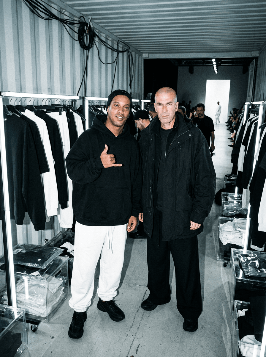 gritty paparazzi-style flash fashion photograph of fashionable subjects standing inside a futuristic backstage styling room filled with illuminated garment rails and transparent acrylic packaging, oversized monochromatic streetwear illuminated by harsh direct flash exposure, chaotic but emotionally detached expressions creating cinematic fashion-week realism
