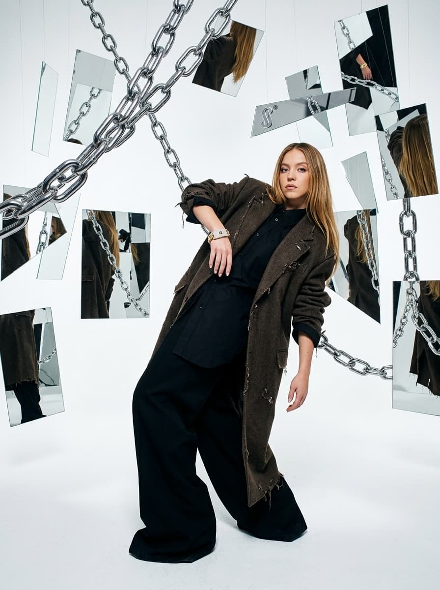 high-fashion studio photograph of a stylish subject wearing the uploaded jewelry (bracelet) paired with an oversized distressed designer coat and exaggerated luxury tailoring, standing inside a giant seamless white studio surrounded by floating chrome chains and suspended mirrors, awkward editorial posture illuminated by harsh DSLR flash exposure, reflective surfaces creating cinematic luxury campaign realism