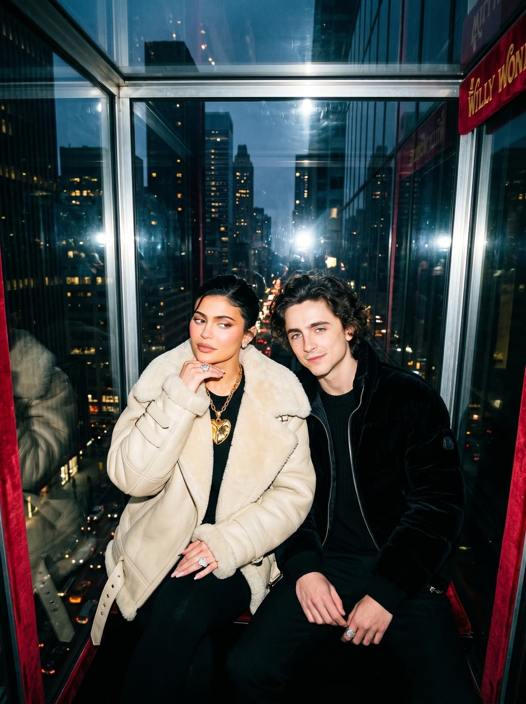aggressive paparazzi-style flash photograph of a fashionable couple wearing oversized luxury outerwear while seated inside a suspended glass cube hanging above a city skyline, dramatic fashion posture illuminated by harsh direct flash lighting, architectural reflections and cinematic luxury campaign atmosphere