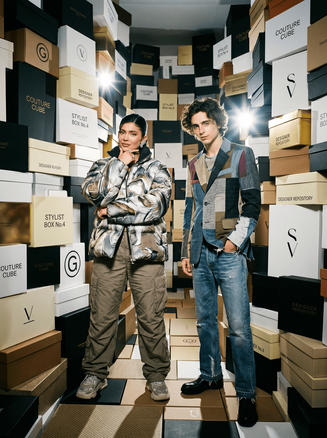 high-fashion studio photograph of a stylish couple wearing coordinated designer fashion while standing inside a giant room made entirely from stacked luxury clothing boxes, awkward editorial body language illuminated by aggressive paparazzi flash exposure, dramatic shadows and product-filled architecture creating cinematic streetwear campaign realism