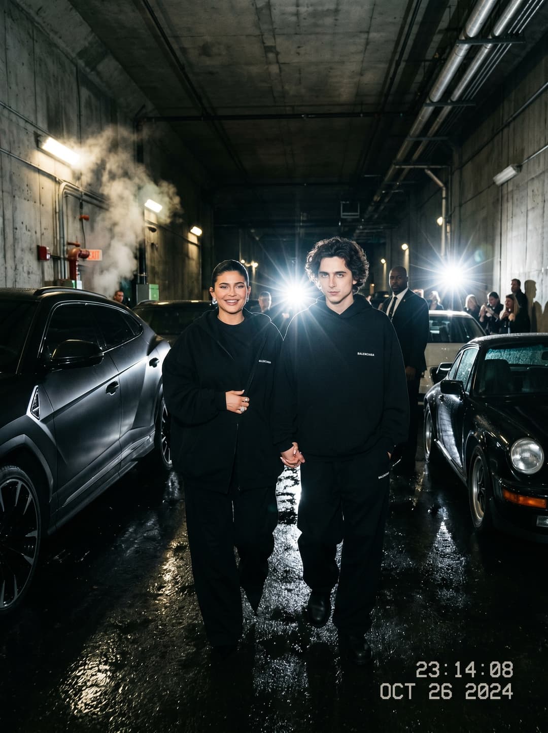 cinematic paparazzi-style flash photograph of a fashionable couple walking through a dark underground tunnel filled with luxury vehicles and reflective wet concrete, oversized monochromatic designer fashion illuminated by aggressive direct flash lighting, antihero energy and cinematic shadows creating luxury Gotham campaign realism