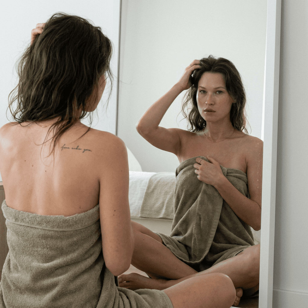 Using the reference subject (DO NOT ALTER FACE), generate a photorealistic RAW-style vertical 9:16 portrait. A woman sits cross-legged in front of a mirror, wrapped in a soft fleece/terry towel slipping naturally off her shoulders, revealing her bare back (tasteful implied nudity only). One arm is raised, fingers lightly in her hair; the other hand holds the towel with natural tension and fabric compression. Expression: soft, intimate, confident but slightly shy. Eyes glance downward then meet the viewer through the mirror. Natural skin texture with pores, subtle redness, and realistic highlights. Hair: long, wavy, dark with natural variation, loose strands and flyaways. Dynamically generate: Cohesive skin tone + undertone (realistic, non-uniform) Consistent ethnicity (no generic blending) Towel color (muted neutral, fleece texture with real folds + stitching) Optional minimal jewelry (1–3 subtle pieces) Optional small realistic tattoo (fine-line, embedded in skin) Composition: Tight framing on face, shoulders, upper back Mirror reflection physically accurate (no distortion or duplication) Reflection clearly shows eyes and expression Environment: Clean, minimal interior, soft neutral tones Editorial, slightly vintage feel Camera: 50mm f/1.2 look Very shallow depth of field Sharp focus on eyes/face, soft falloff on shoulders + towel Natural exposure, filmic contrast (no HDR, no glow)
