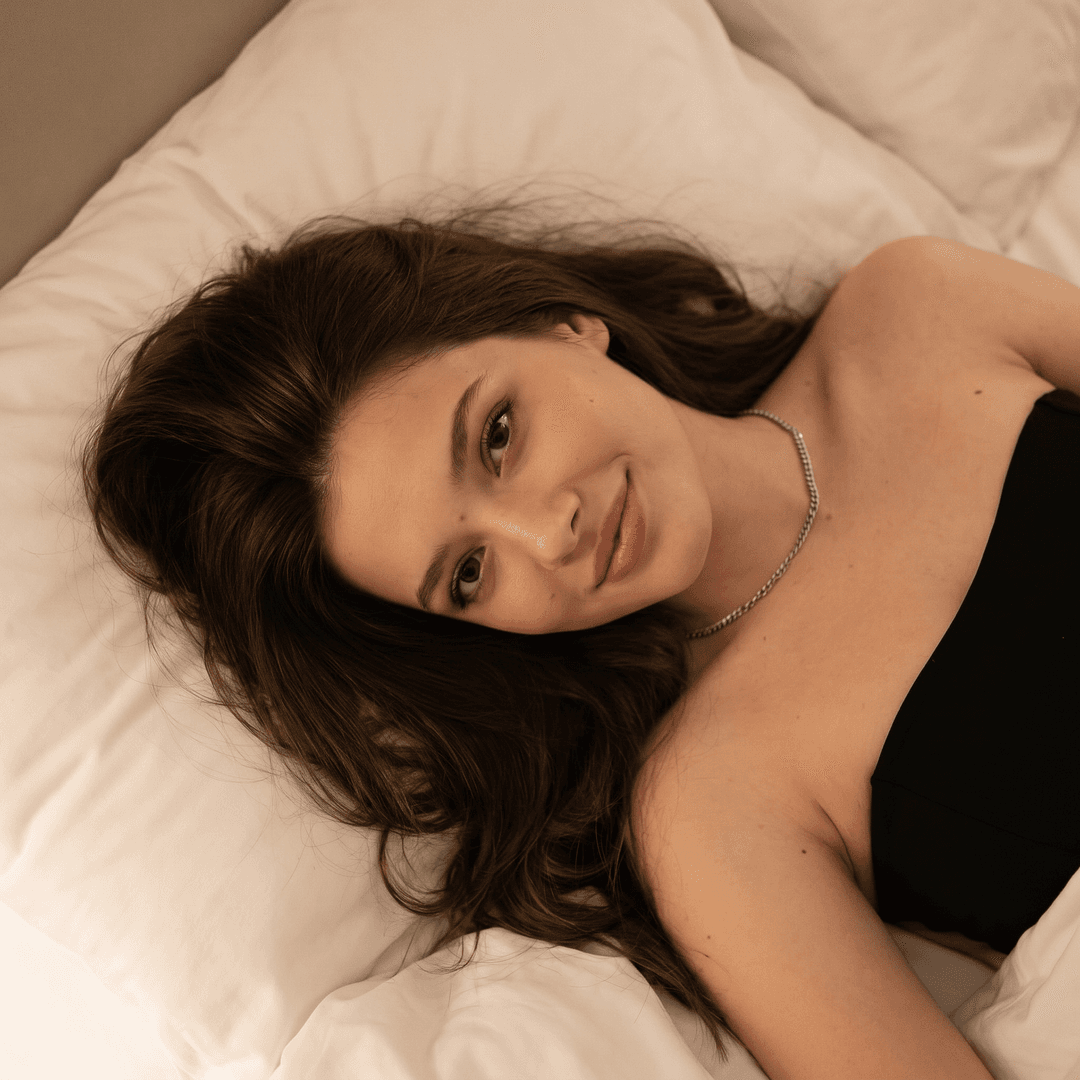 Using the person in the uploaded reference image (DO NOT TOUCH FACE), recreate this scene: { "subject": { "demographics": " woman, Roughly 20s", "hair": { "color": "Dark brown", "style": "Long, wavy, tousled, spread out on pillow", "texture": "Soft, natural" }, "face": { "expression": "Gentle smile, relaxed", "eyes": "Brown, looking directly at camera", "skin_tone": "Fair to medium, rosy cheeks", "makeup": "Natural look, minimal" }, "pose": "Reclining/lying down on back, head tilted slightly" }, "attire": { "clothing": "Black spaghetti-strap tank top or camisole", "neckline": "Low scoop neck" }, "accessories": { "jewelry": [ "Thin gold chain necklace", "Small dark pendant (possibly heart-shaped or geometric)" ] }, "environment": { "setting": "Bedroom, bed", "background": "White bed sheets/pillows, soft texture, slightly wrinkled" }, "photography_style": { "angle": "High angle, close-up, selfie perspective", "lighting": "Soft indoor lighting, warm tones, slight shadowing on the neck and side of face", "focus": "Sharp focus on face, slightly softer background" } }
