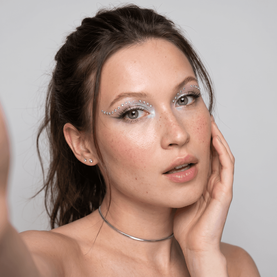 Using the person in the uploaded reference image, recreate this scene: ultra-realistic beauty selfie portrait of the person from the reference image, captured at a slight three-quarter angle, close-up framing from shoulders up. The camera is held in hand, selfie perspective. Facial expression: mouth slightly open with relaxed parted lips, soft sensual expression, subtle surprise or playful curiosity in the eyes. Extremely realistic skin texture with visible pores, soft freckles, natural blush, and healthy glow. Dramatic eye makeup with shimmering pastel tones, precise eyeliner, long lashes and decorative rhinestone makeup forming symmetrical curved lines. Hairstyle with loose front strands and ponytail. Wearing small stud earrings and a thin choker necklace. Soft even beauty studio-like lighting. Clean neutral background. Ultra high resolution, 8K, HD detail, razor-sharp focus, professional photography look.

