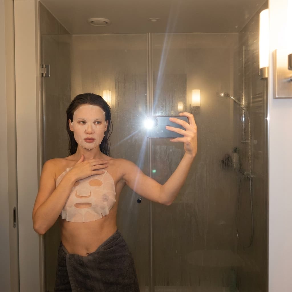 A medium shot captures the person in the uploaded reference image (DO NOT TOUCH FACE) taking a selfie in a steamy bathroom mirror. The person's torso is visible from the chest down, with a white sheet mask covering their chest and a dark grey towel wrapped around their waist. Their left arm is extended, holding a smartphone that is capturing the reflection. The right hand is placed on top of the sheet mask. The background shows a shower stall with a glass door covered in condensation, and two wall-mounted light fixtures above. The lighting is soft and warm, emanating from the phone's flash and the bathroom lights, creating a slight lens flare. The camera is positioned at eye level, with a slightly 24-28mm equivalent lens perspective. The overall tone is intimate and casual.
