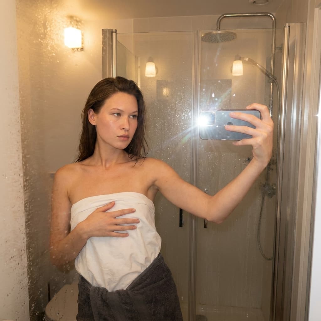 A medium shot captures the person in the uploaded reference image (DO NOT TOUCH FACE) taking a selfie in a steamy bathroom mirror. The person's torso is visible from the chest down, with a white sheet covering their chest and a dark grey towel wrapped around their waist. Their left arm is extended, holding a smartphone that is capturing the reflection. The right hand is placed on top of the sheet. The background shows a shower stall with a glass door covered in condensation, and two wall-mounted light fixtures above. The lighting is soft and warm, emanating from the phone's flash and the bathroom lights, creating a slight lens flare. The camera is positioned at eye level, with a slightly 24-28mm equivalent lens perspective. The overall tone is intimate and casual.