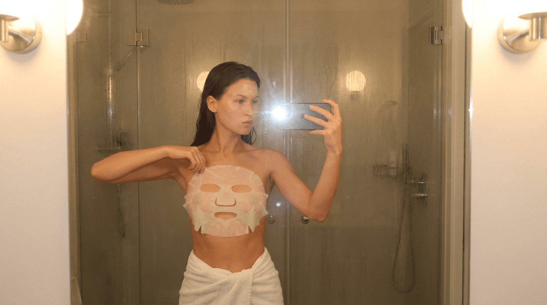 A medium shot captures the person in the uploaded reference image (DO NOT TOUCH FACE) taking a selfie in a steamy bathroom mirror. The person's torso is visible from the chest down, with a white sheet mask covering their chest and a a white towel wrapped around their waist. Their left arm is extended, holding a smartphone that is capturing the reflection. The right hand is placed on neck of the sheet mask. The background shows a shower stall with a glass door covered in condensation, and two wall-mounted light fixtures above. The lighting is soft and warm, emanating from the phone's flash and the bathroom lights, creating a slight lens flare. The camera is positioned at eye level, with a slightly 24-28mm equivalent lens perspective. The overall tone is intimate and casual.