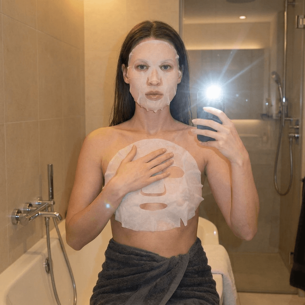 A medium shot captures the person in the uploaded reference image (DO NOT TOUCH FACE). The person's torso is visible from the chest down, with a white sheet mask covering their chest and a dark grey towel wrapped around their waist. The right hand is placed on top of the sheet mask. The lighting is soft and warm, emanating from the phone's flash and the bathroom lights, creating a slight lens flare. The camera is positioned at eye level, with a slightly 24-28mm equivalent lens perspective. The overall tone is intimate and casual.