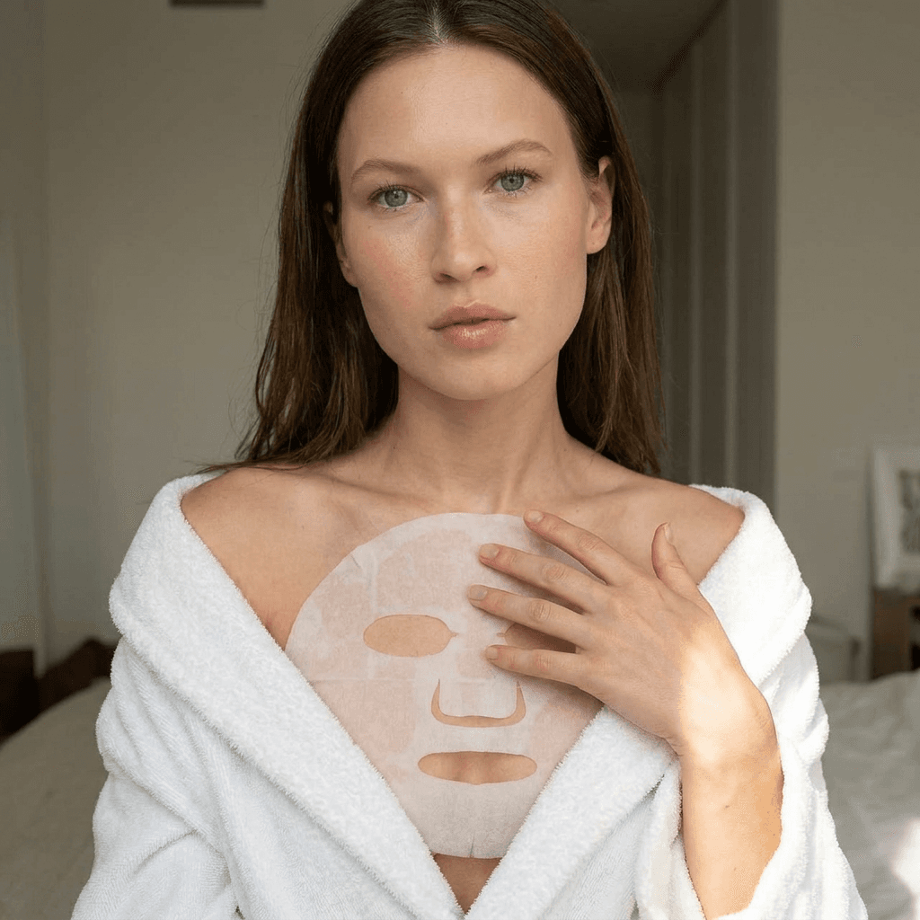 A medium shot captures the person in the uploaded reference image (DO NOT TOUCH FACE). The person's torso is visible from the chest down, with a white sheet mask covering their chest. The right hand is placed on top of the sheet mask. The lighting is soft and warm. The camera is positioned at eye level, with a slightly 24-28mm equivalent lens perspective. The overall tone is intimate and casual.