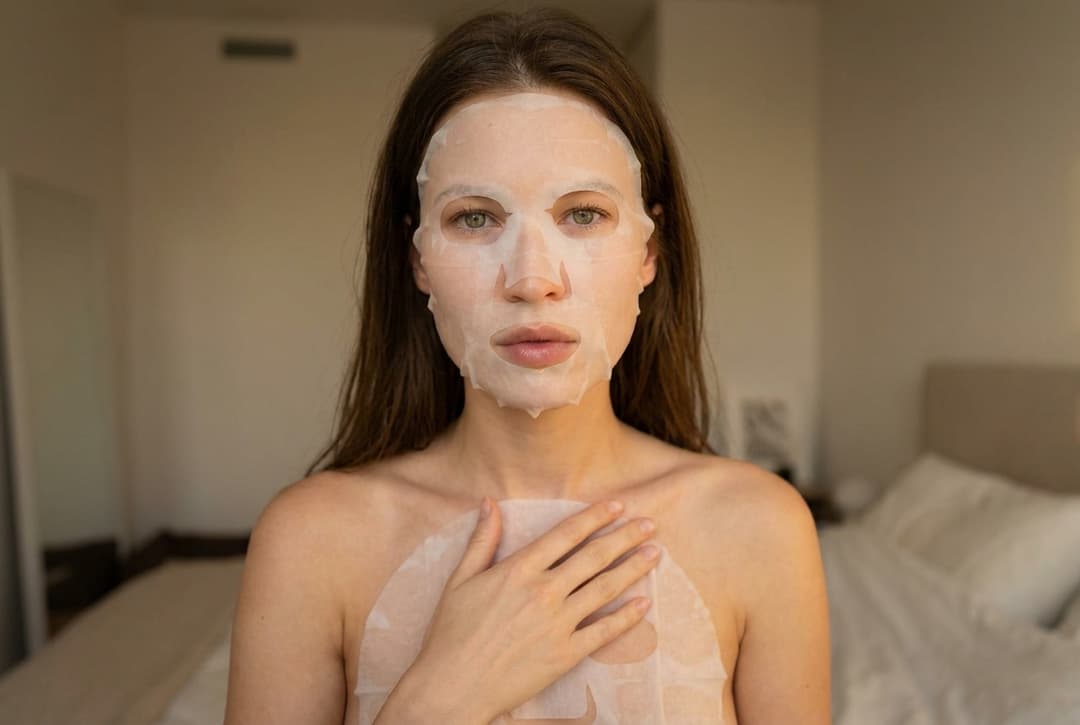 A medium shot captures the person in the uploaded reference image (DO NOT TOUCH FACE). no robe. The person's torso is visible from the chest down, with a white invisible mask covering their chest. The right hand is placed on top of the sheet mask. The lighting is soft and warm. The camera is positioned at eye level, with a slightly 24-28mm equivalent lens perspective. The overall tone is intimate and casual.