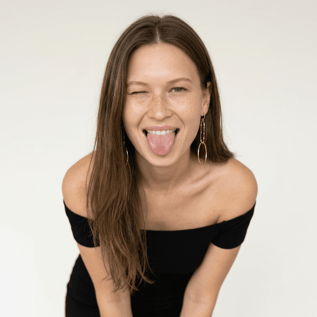 Using the person in the uploaded reference image, recreate this scene: { "style": "ultra-realistic studio portrait", "subject": { "gender": "female", "age": " ", "pose": "leaning slightly forward toward the camera", "expression": "playful, flirty", "facial_details": { "wink": true, "tongue_out": true, "freckles": "natural across fair skin", "makeup": { none } }, "hair": { "color": "", "length": "long", "part": "side-parted", "style": "falling naturally over shoulders" }, "outfit": { "dress": "off-shoulder fitted dress", "jewelry": { "earrings": "long dangling gold earrings" } } }, "environment": { "setting": "studio", "background": "clean minimal light neutral tones" }, "lighting": { "type": "soft diffused studio lighting", "shadows": "smooth natural shadows" }, "camera": { "lens": "50mm", "aperture": "f/1.8", "depth_of_field": "shallow" }, "quality": { "resolution": "high resolution", "detail": "ultra-detailed", "skin_texture": "photorealistic", "focus": "sharp focus", "photography_style": "high fashion lifestyle photography" } }
