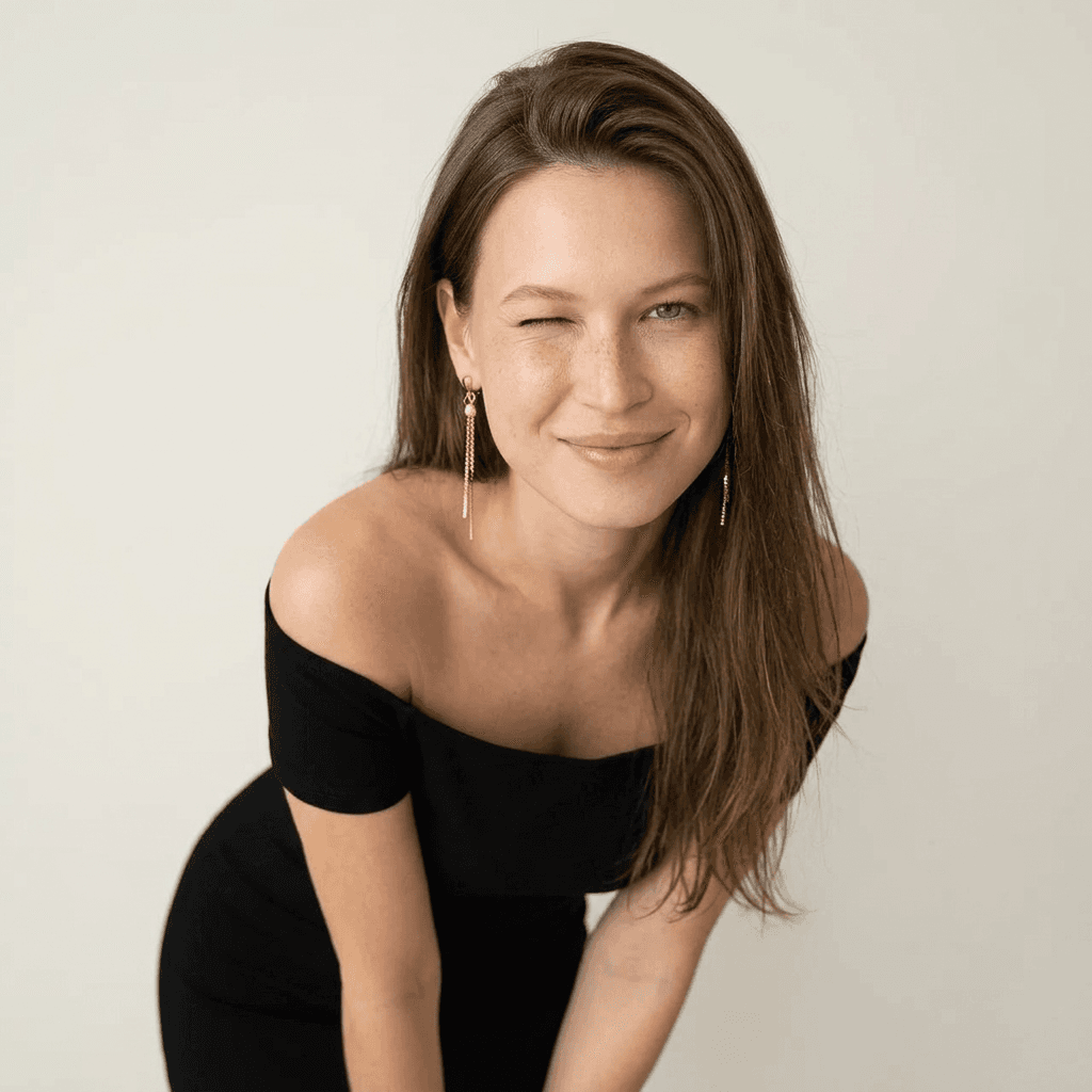 Using the person in the uploaded reference image, recreate this scene: { "style": "ultra-realistic studio portrait", "subject": { "gender": "female", "age": " ", "pose": "leaning slightly forward toward the camera", "expression": "playful, flirty", "facial_details": { "wink": true, : true, "freckles": "natural across fair skin", "makeup": { none } }, "hair": { "color": "", "length": "long", "part": "side-parted", "style": "falling naturally over shoulders" }, "outfit": { "dress": "off-shoulder fitted dress", "jewelry": { "earrings": "long dangling gold earrings" } } }, "environment": { "setting": "studio", "background": "clean minimal light neutral tones" }, "lighting": { "type": "soft diffused studio lighting", "shadows": "smooth natural shadows" }, "camera": { "lens": "50mm", "aperture": "f/1.8", "depth_of_field": "shallow" }, "quality": { "resolution": "high resolution", "detail": "ultra-detailed", "skin_texture": "photorealistic", "focus": "sharp focus", "photography_style": "high fashion lifestyle photography" } }
