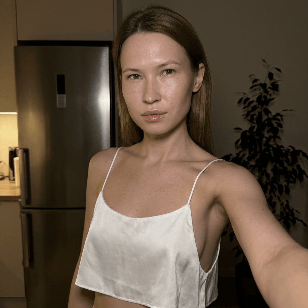 Using the person in the uploaded reference image (DO NOT TOUCH FACE), recreate this scene: Ультрареалистичный селфи-снимок в условиях глубокой ночи. Мягкое, приглушённое освещение, тёплый слабый источник света сбоку (например, от ночника или кухни), создаёт спокойную, интимную атмосферу. Без вспышки. В кадре девушка: прямые волосы чуть ниже плечь, закинуты назад. смотрит в камеру. Молодое лицо. Кожа естественного, реалистичного оттенка, лёгкий блеск на губах. Ракурс — слегка сбоку, как будто фото сделано одной рукой. Фон: часть серебристого холодильника, рядом заметен высокий декоративный цветок, слегка в тени. Одежда: свободная, белая укороченная атласная майка из тончайшей ткани на тонких бретелях, с экстремально глубокими вырезами в зоне подмышек. Атмосфера спокойная, домашняя, ночная, с мягкими тенями и приглушёнными цветами. Формат 9:16. Photorealistic capture with iPhone 15 Pro style optics, natural skin texture, realistic reflections, balanced exposure, and sharp focus.