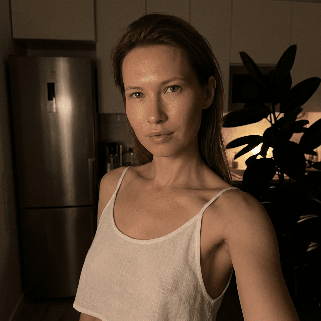 Using the person in the uploaded reference image (DO NOT TOUCH FACE), recreate this scene: Ультрареалистичный селфи-снимок в условиях глубокой ночи. Мягкое, приглушённое освещение, тёплый слабый источник света сбоку (например, от ночника или кухни), создаёт спокойную, интимную атмосферу. Без вспышки. В кадре девушка: прямые волосы чуть ниже плечь, закинуты назад. смотрит в камеру. Молодое лицо. Кожа естественного, реалистичного оттенка, лёгкий блеск на губах. Ракурс — слегка сбоку, как будто фото сделано одной рукой. Фон: часть серебристого холодильника, рядом заметен высокий декоративный цветок, слегка в тени. Одежда: свободная, белая укороченная майка из очень тонкой, прозрачной ткани на тонких бретелях, с экстремально глубокими вырезами в зоне подмышек. Атмосфера спокойная, домашняя, ночная, с мягкими тенями и приглушёнными цветами. Формат 9:16. Photorealistic capture with iPhone 15 Pro style optics, natural skin texture, realistic reflections, balanced exposure, and sharp focus.