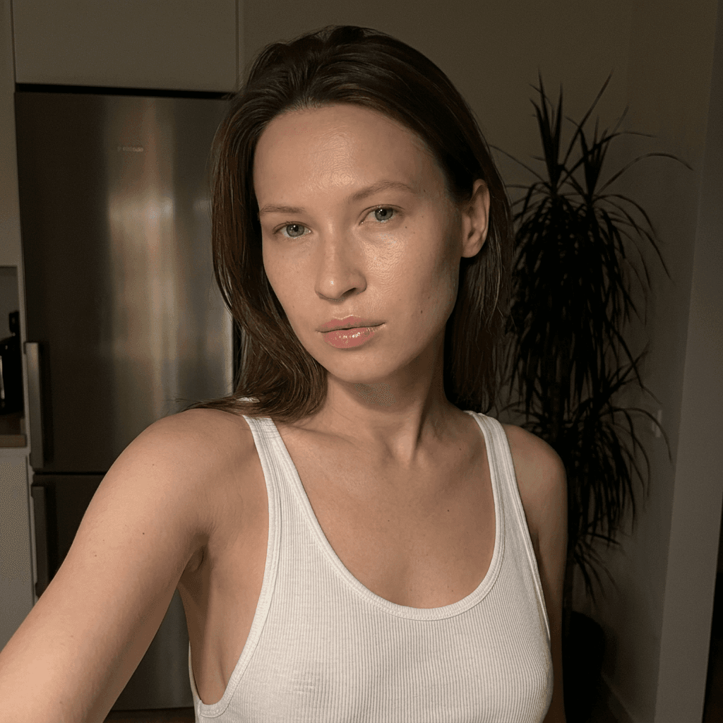 Using the person in the uploaded reference image (DO NOT TOUCH FACE), recreate this scene: Ультрареалистичный селфи-снимок в условиях глубокой ночи. Мягкое, приглушённое освещение, тёплый слабый источник света сбоку (например, от ночника или кухни), создаёт спокойную, интимную атмосферу. Без вспышки. В кадре девушка: прямые волосы чуть ниже плечь, закинуты назад. смотрит в камеру. Молодое лицо. Кожа естественного, реалистичного оттенка, лёгкий блеск на губах. Ракурс — слегка сбоку, как будто фото сделано одной рукой. Фон: часть серебристого холодильника, рядом заметен высокий декоративный цветок, слегка в тени. Одежда: свободная, белая укороченная майка из очень тонкой, прозрачной ткани на тонких бретелях, с экстремально глубокими вырезами в зоне подмышек. Атмосфера спокойная, домашняя, ночная, с мягкими тенями и приглушёнными цветами. Формат 9:16. Photorealistic capture with iPhone 15 Pro style optics, natural skin texture, realistic reflections, balanced exposure, and sharp focus.