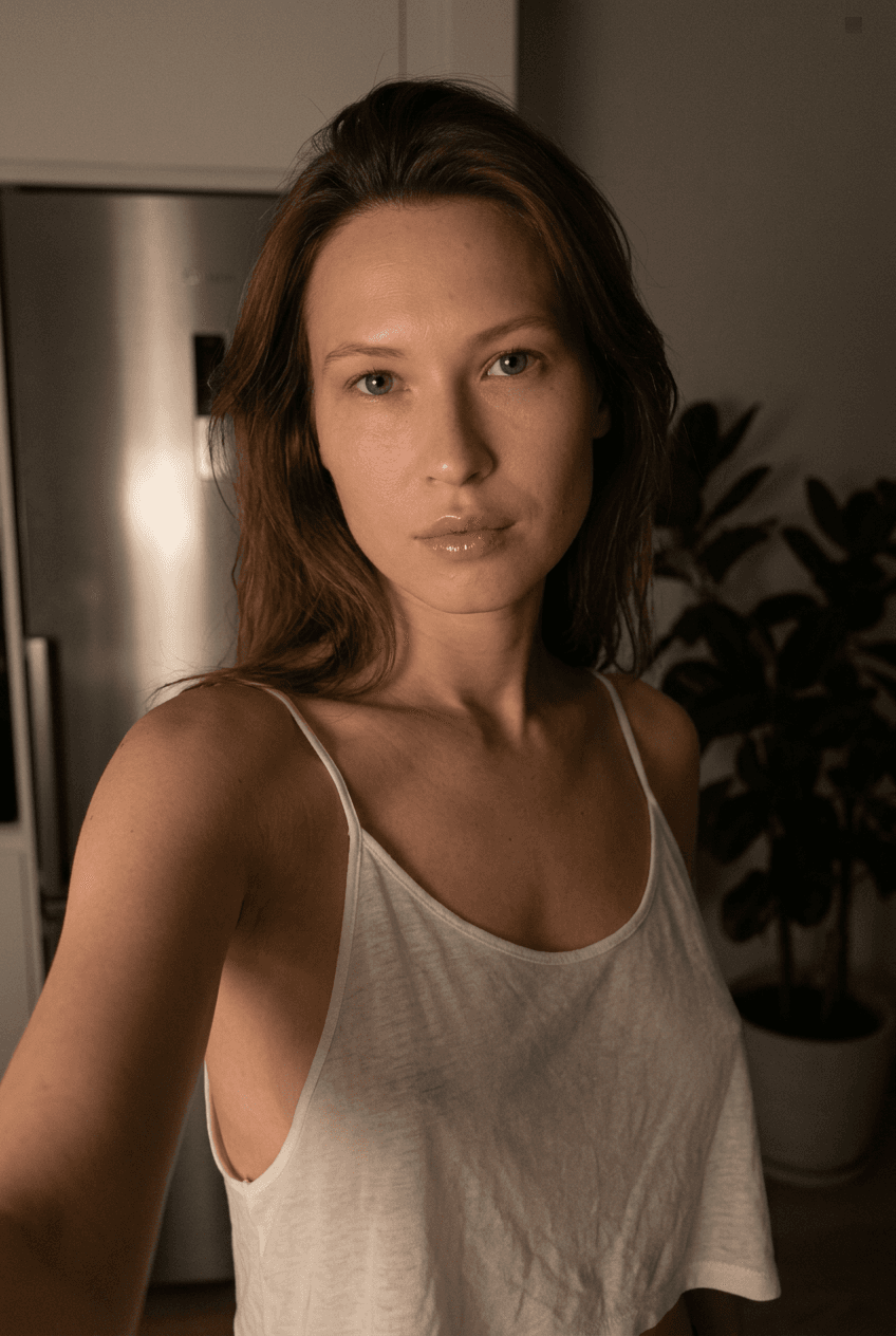 Using the person in the uploaded reference image (DO NOT TOUCH FACE), recreate this scene: Ультрареалистичный селфи-снимок в условиях глубокой ночи. Мягкое, приглушённое освещение, тёплый слабый источник света сбоку (например, от ночника или кухни), создаёт спокойную, интимную атмосферу. Без вспышки. В кадре девушка: прямые волосы чуть ниже плечь, закинуты назад. смотрит в камеру. Молодое лицо. Кожа естественного, реалистичного оттенка, лёгкий блеск на губах. Ракурс — слегка сбоку, как будто фото сделано одной рукой. Фон: часть серебристого холодильника, рядом заметен высокий декоративный цветок, слегка в тени. Одежда: свободная, сильно укороченная майка из очень тонкой, почти невидимой бело-прозрачной ткани на тонких бретелях, с экстремально глубокими вырезами в зоне подмышек. Атмосфера спокойная, домашняя, ночная, с мягкими тенями и приглушёнными цветами. Формат 9:16. Photorealistic capture with iPhone 15 Pro style optics, natural skin texture, realistic reflections, balanced exposure, and sharp focus.