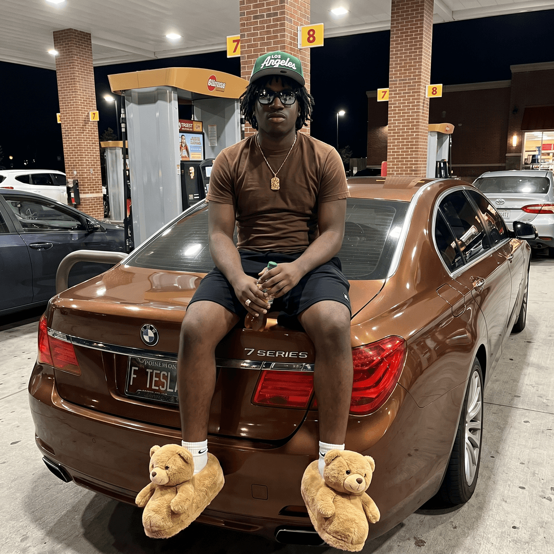 ABSOLUTE photorealism, real-life street photography, no cartoon or CGI appearance. Ultra‑realistic candid nighttime photo of a young man sitting confidently on the trunk of a luxury brown BMW sedan at a gas station. The subject has a relaxed, stylish, modern streetwear vibe. He wears a green Los Angeles baseball cap, dark reflective sunglasses, a brown fitted T‑shirt, black athletic shorts, and oversized plush teddy‑bear slippers. Medium-dark skin tone, youthful face, natural posture, authentic expression, realistic fabric textures, accurate wrinkles, and subtle jewelry. The brown BMW sedan features glossy paint, chrome accents, realistic reflections, and a custom license plate reading “F TESLA,” with the trunk surface catching the bright overhead gas station lights. The environment includes brick columns, fuel pumps labeled 7 and 8, concrete ground with subtle texture, and warm nighttime ambience. Cinematic lighting with mixed warm and cool tones, soft reflections on the car, and realistic shadows. Shot style resembles a real candid iPhone 17 Pro night photo with ultra‑realistic low-light rendering, natural body language, authentic facial expression, subtle motion blur, realistic flash reflections, cinematic depth of field, and magazine-level street photography realism. High-contrast nighttime lighting, 8K hyper-detail, luxury car culture energy, effortlessly cool streetwear moment captured with authentic real-world atmosphere.

[Character "Jesus": A teenage boy stands in a bright outdoor corridor with a white textured wall behind him and palm trees visible in the background. He has a medium‑brown skin tone, a youthful round face, and wears rectangular glasses with dark frames. His hair is short, dark, and neatly trimmed. He has a relaxed, confident posture, standing casually with one hand holding a plastic bottle filled with a brown liquid.

He wears a white graphic T‑shirt featuring a large colorful illustrated face and bold stylized text. He pairs it with black athletic shorts that fall above the knee. On his feet are white crew socks and brown patterned slide sandals. A thin gold chain necklace rests around his neck, adding a subtle touch of style. The lighting is natural daylight, giving the scene a warm, tropical feel.]