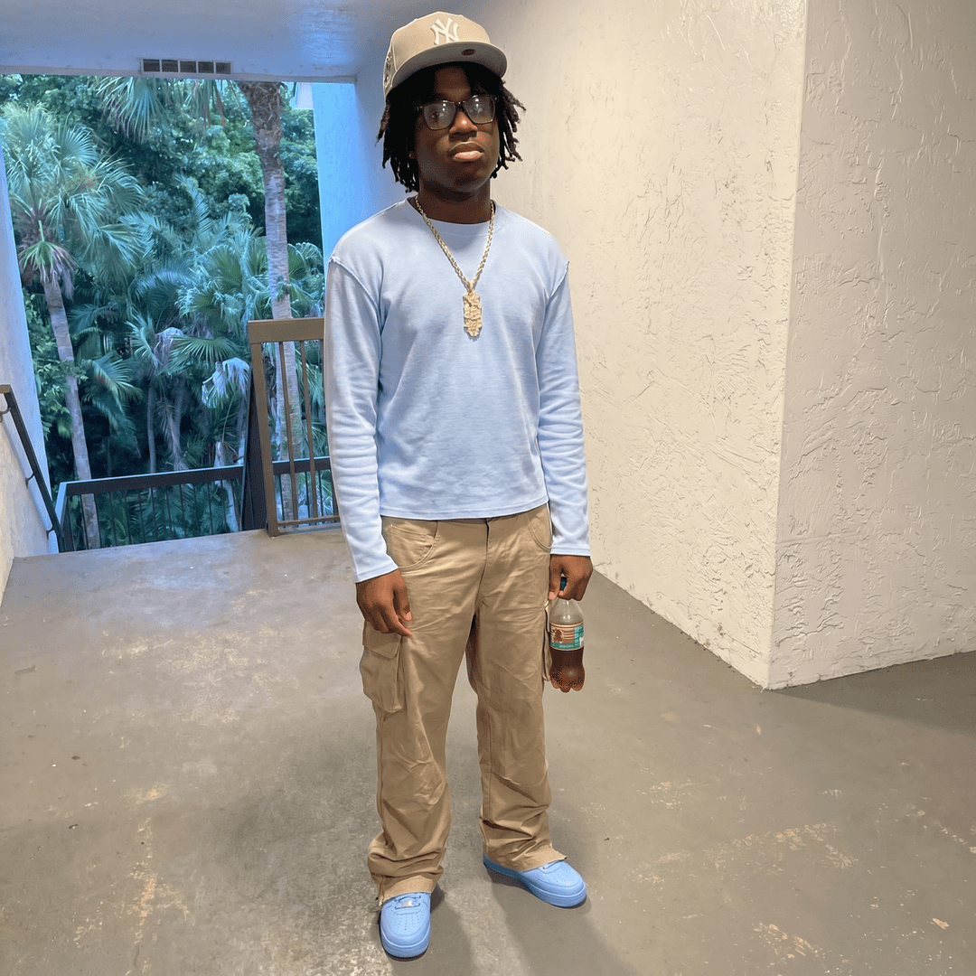put me instead of him but put his fit on me

[Character "Jesus": A teenage boy stands in a bright outdoor corridor with a white textured wall behind him and palm trees visible in the background. He has a medium‑brown skin tone, a youthful round face, and wears rectangular glasses with dark frames. His hair is short, dark, and neatly trimmed. He has a relaxed, confident posture, standing casually with one hand holding a plastic bottle filled with a brown liquid.

He wears a white graphic T‑shirt featuring a large colorful illustrated face and bold stylized text. He pairs it with black athletic shorts that fall above the knee. On his feet are white crew socks and brown patterned slide sandals. A thin gold chain necklace rests around his neck, adding a subtle touch of style. The lighting is natural daylight, giving the scene a warm, tropical feel.]