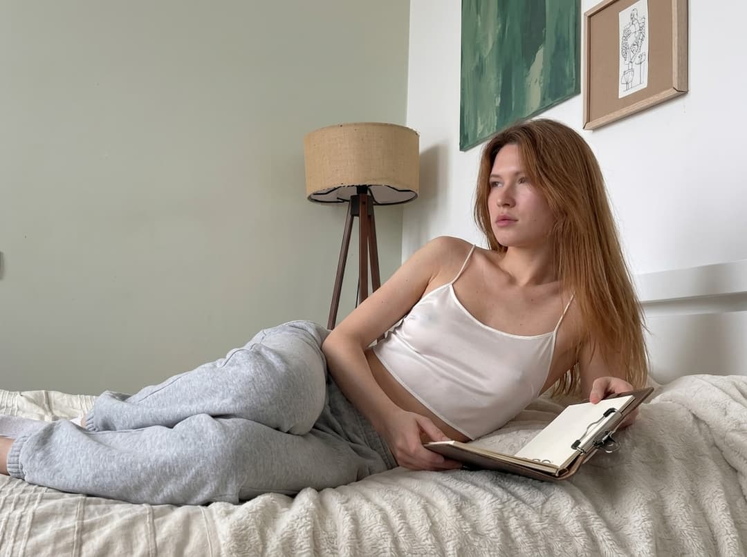 “A medium shot captures the person in the uploaded reference image (DO NOT TOUCH FACE). Одежда: свободная сильно укороченная атласная майка на тонких бретелях из очень тонкой прозрачной ткани как чулки, с глубокими вырезами в зоне подмышек. no covering. The lighting is soft and warm, with a slightly 24-28mm equivalent lens perspective. The overall tone is intimate and casual”