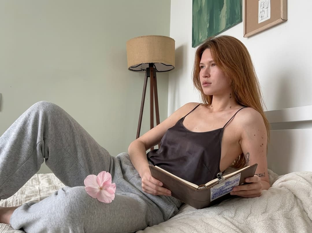 “A medium shot captures the person in the uploaded reference image (DO NOT TOUCH FACE). Одежда: свободная сильно укороченная атласная майка на тонких бретелях из очень тонкой прозрачной темной ткани, с глубокими вырезами в зоне подмышек. no covering. The lighting is soft and warm, with a slightly 24-28mm equivalent lens perspective. The overall tone is intimate and casual”